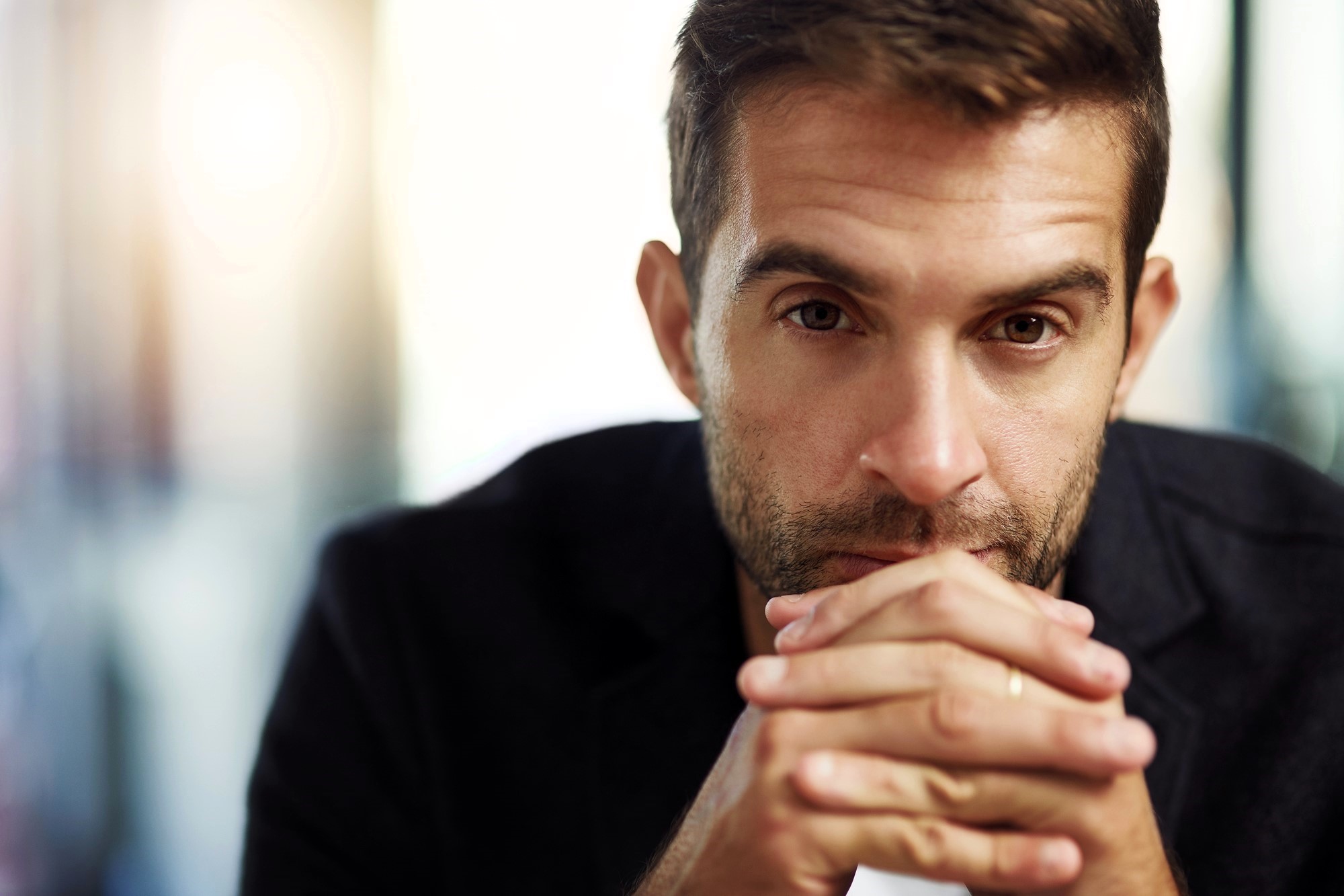 A man with short brown hair and a beard gazes intensely at the camera, resting his hands under his chin. He wears a dark jacket, and the background is softly blurred, creating a focused portrait.