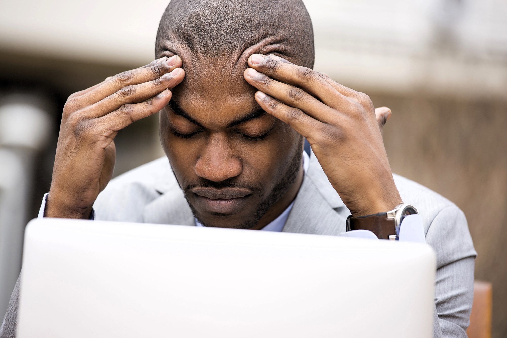 A man in a suit sits in front of a laptop, resting his forehead in his hands. His eyes are closed, and he appears deep in thought or stressed. The background is blurred and neutral.