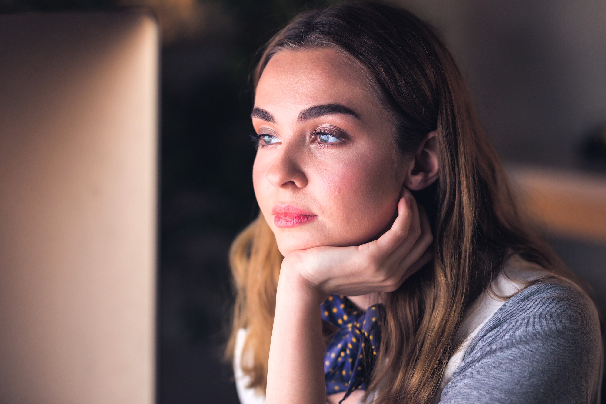 A woman with long brown hair and a thoughtful expression rests her chin on her hand while looking into the distance. She's wearing a gray top and a blue scarf with yellow dots. The background is softly blurred.