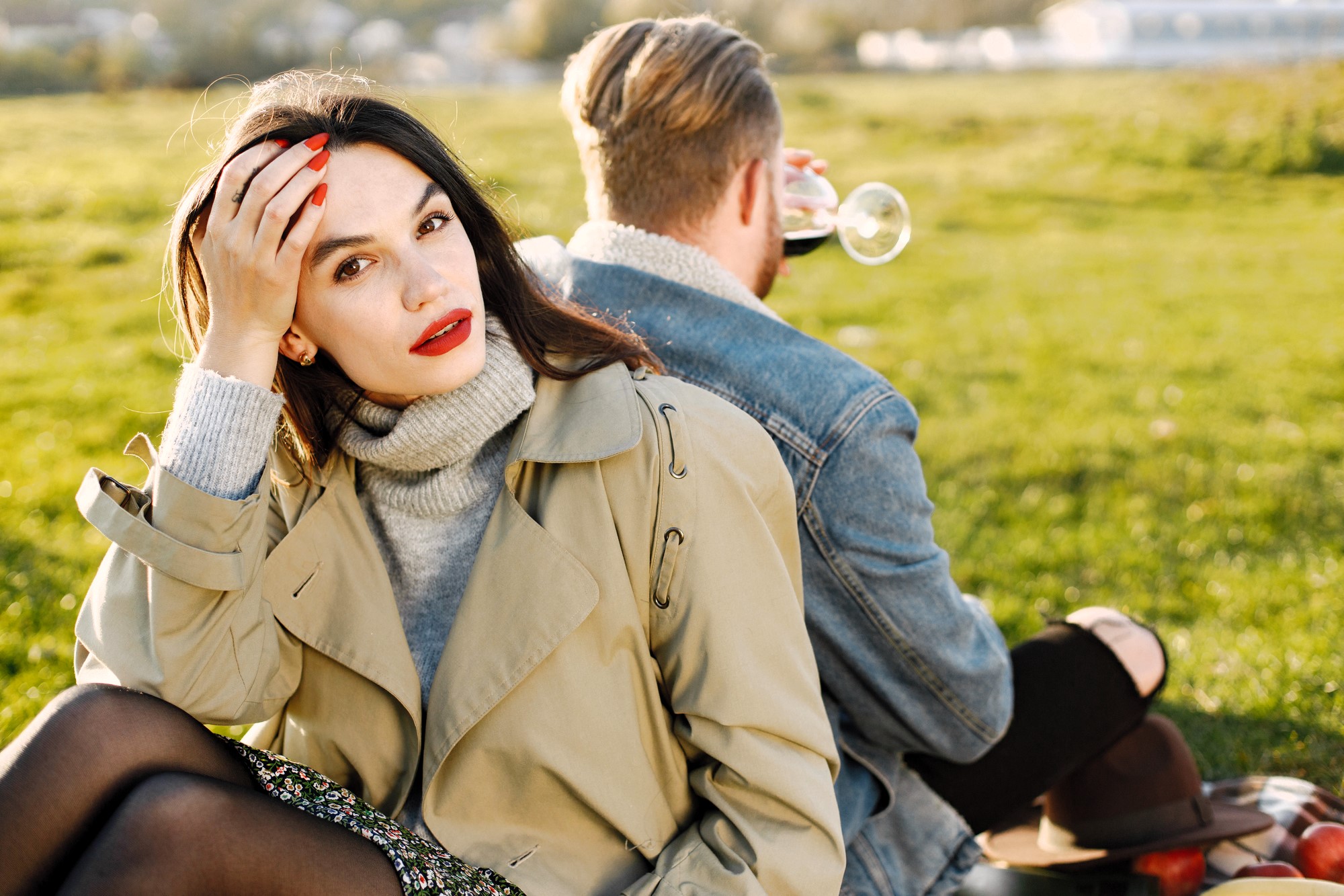 A woman in a beige coat sits on grass, looking at the camera with her hand in her hair. She has red lipstick and painted nails. A man, facing away, drinks from a glass. They are outdoors on a sunny day with blurry greenery in the background.