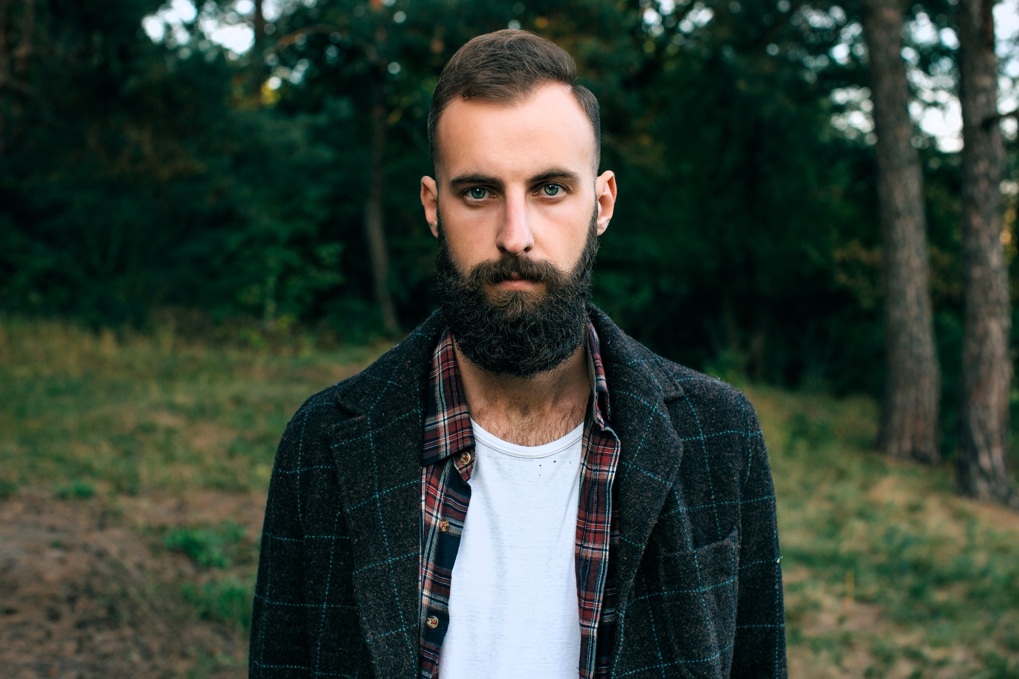 A bearded man stands outdoors in a forested area. He is wearing a dark checkered jacket over a red plaid shirt and a white t-shirt. Tall trees are visible in the background.