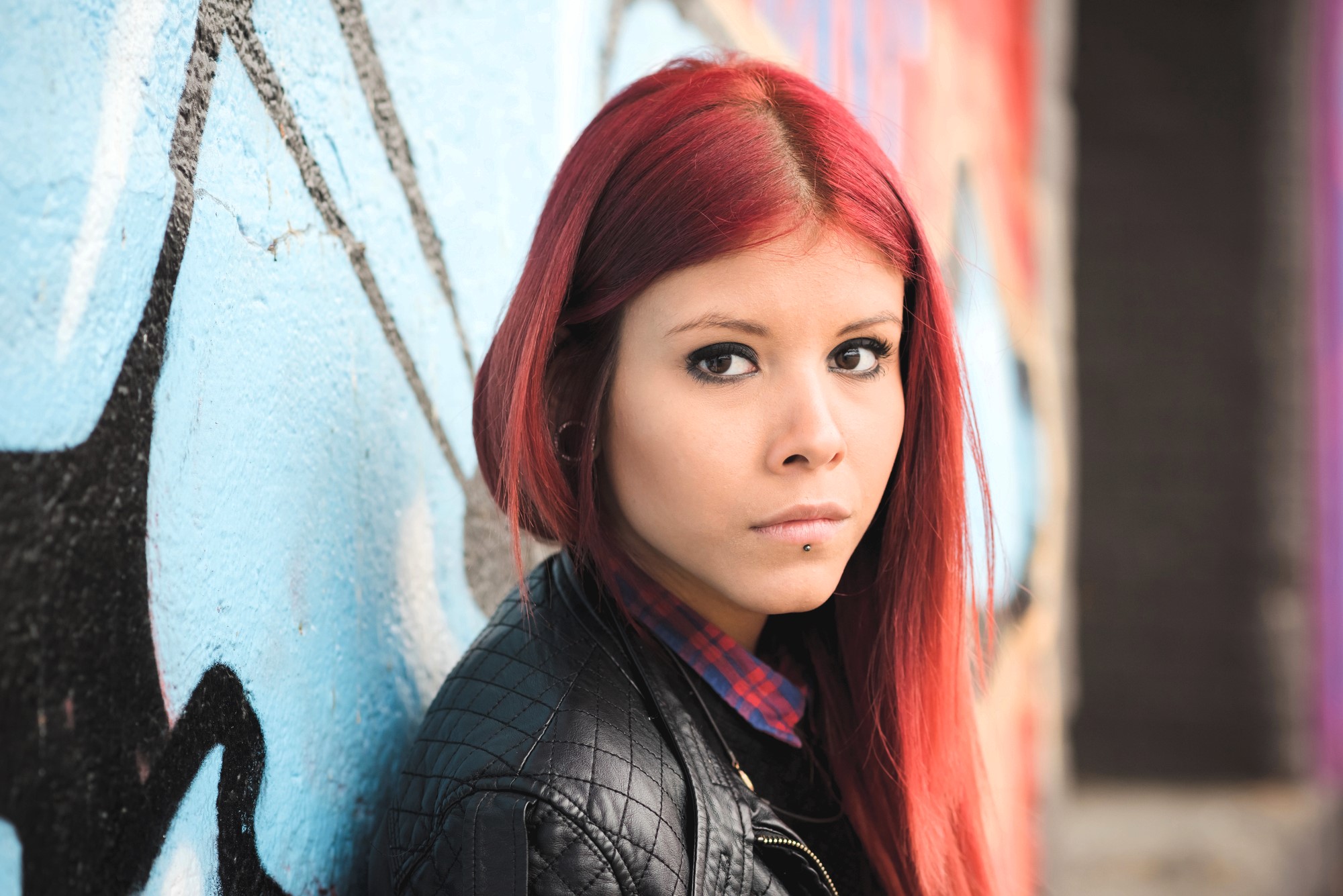 A person with long red hair stands in front of a graffiti-covered wall. They are wearing a black leather jacket and a plaid shirt, looking directly at the camera with a neutral expression.