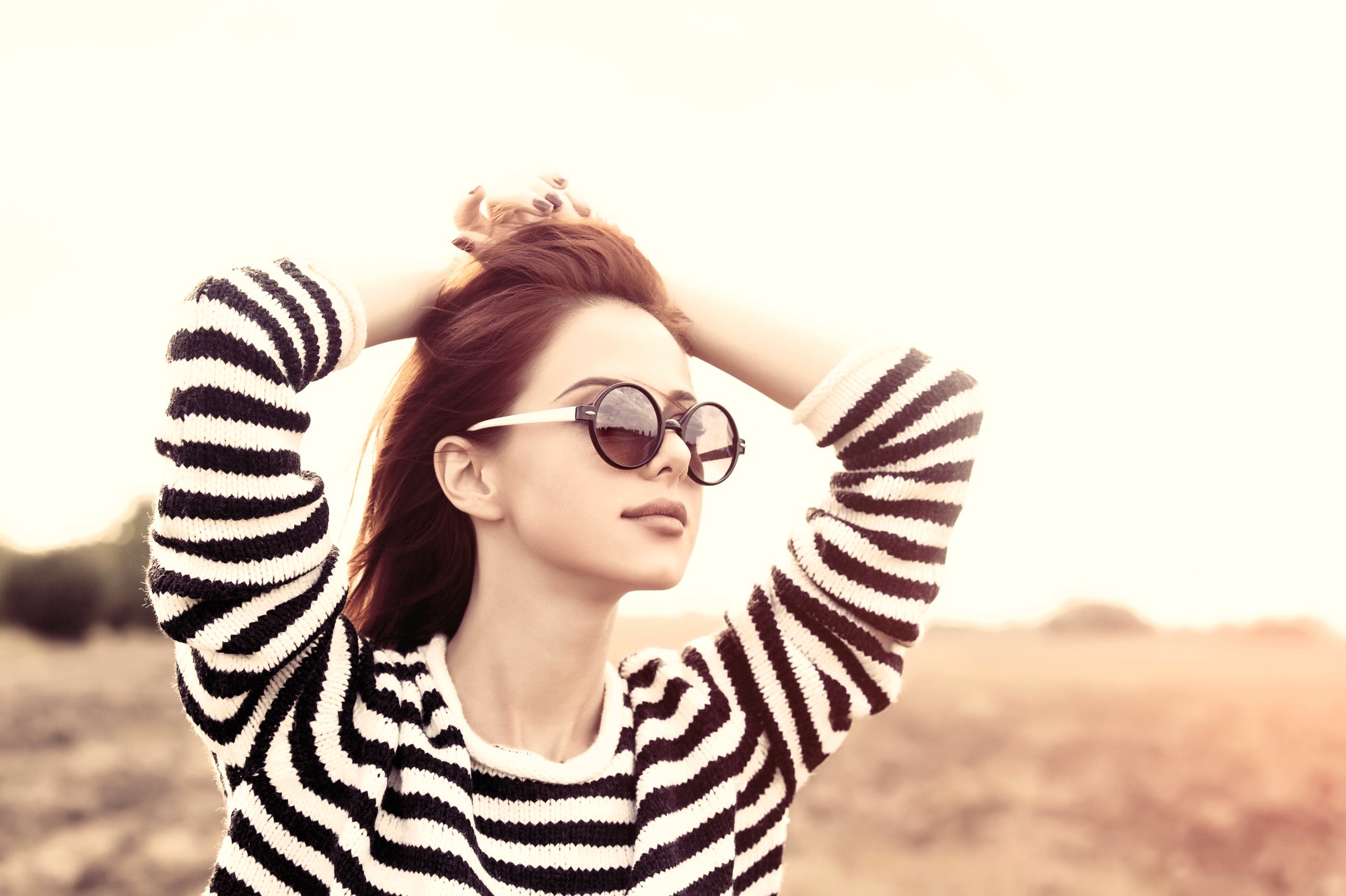 Woman in a striped sweater wearing sunglasses, standing outdoors with hands in her hair. The background features a soft, blurred landscape and a warm, sunlit ambiance.
