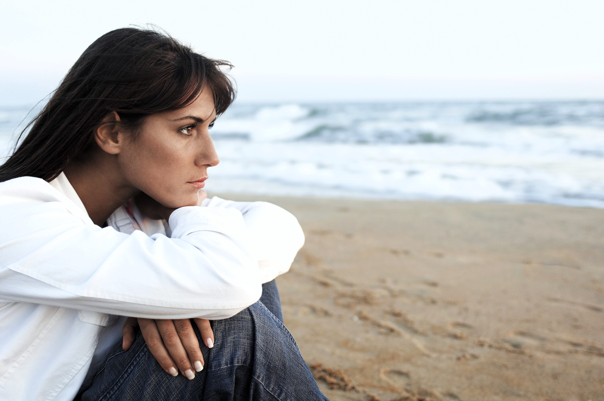 A woman in a white shirt and jeans sits on a sandy beach, gazing thoughtfully towards the sea. The ocean waves are gently rolling in under a cloudy sky. Her long dark hair is flowing freely in the breeze.