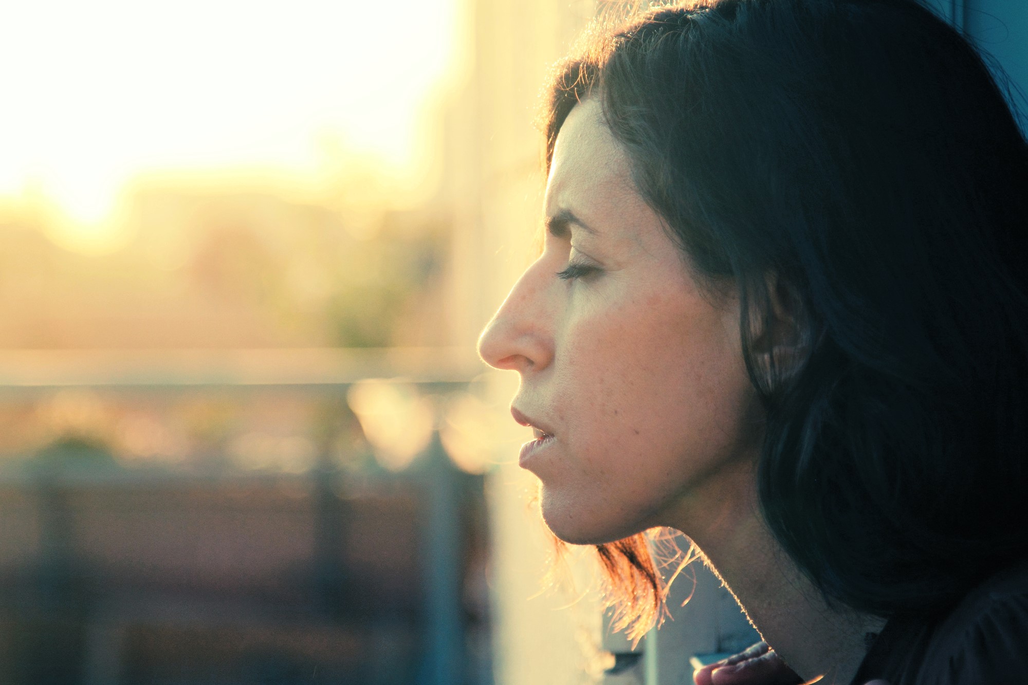 Side profile of a woman with shoulder-length dark hair, eyes closed, basking in the warm glow of sunlight. The background is softly blurred, giving a tranquil, contemplative atmosphere.