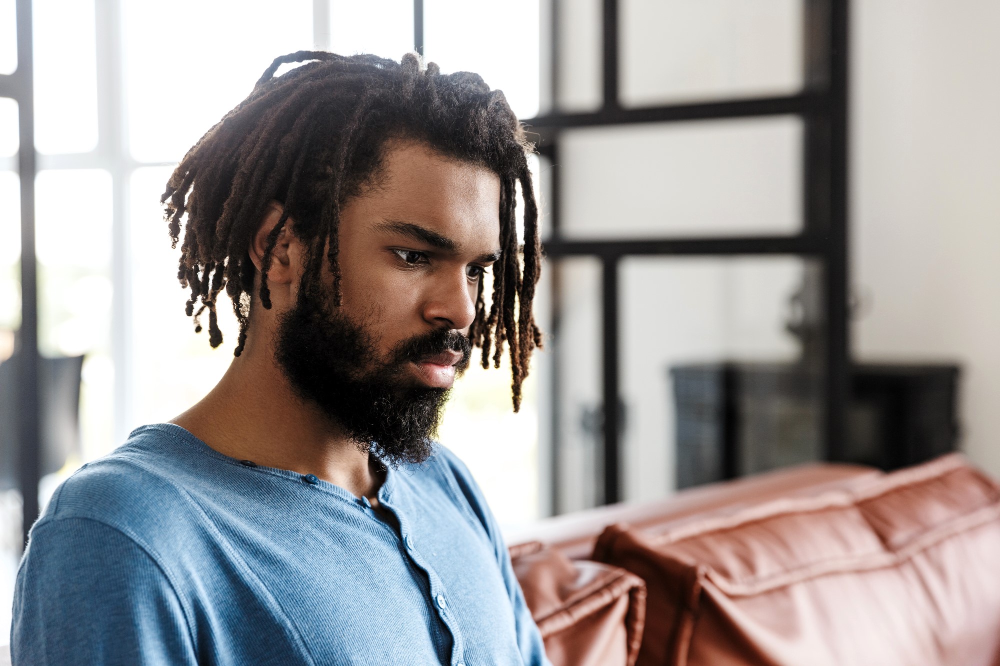 A man with dreadlocks and a beard wearing a blue shirt is sitting on a sofa, looking thoughtful. The room has large windows and natural light.