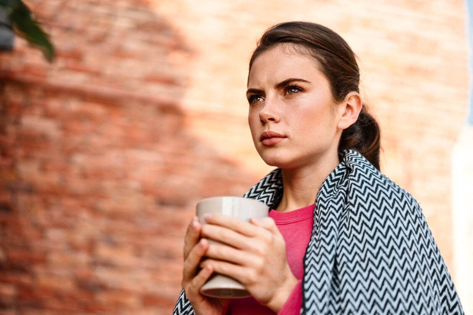 brunette woman with a somewhat serious face holding a hot drink in two hands with a blanket wrapped around her shoulders