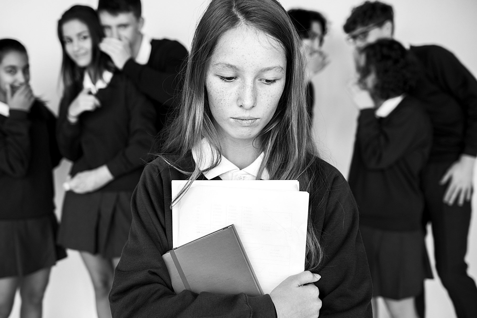 A girl stands alone, looking down and holding books. In the background, a group of students in uniforms are whispering and pointing, creating a sense of exclusion. The image is in black and white.