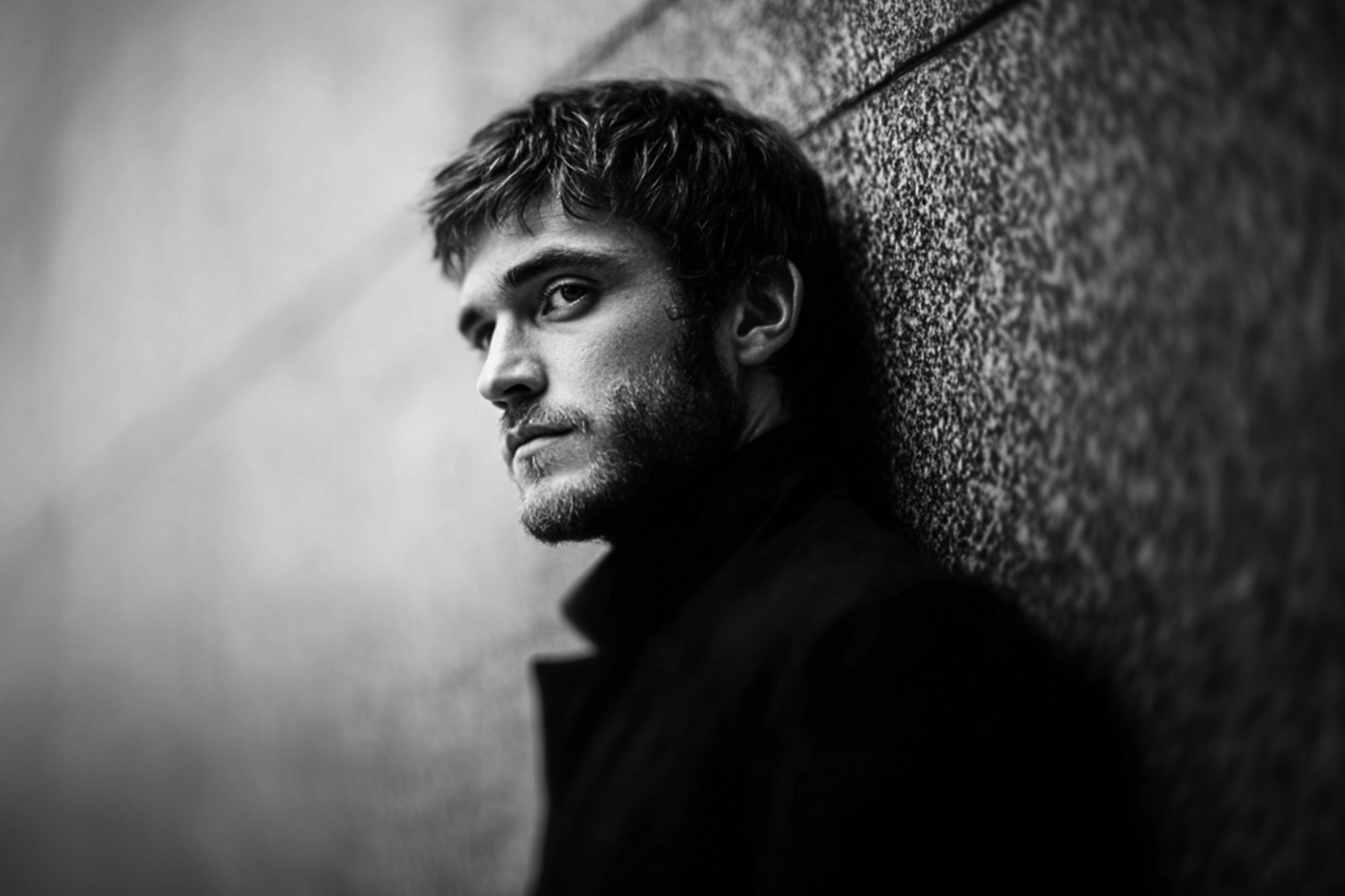A man with short hair and a beard leans against a textured wall, gazing intensely at the camera. The black and white image creates a dramatic and moody atmosphere.