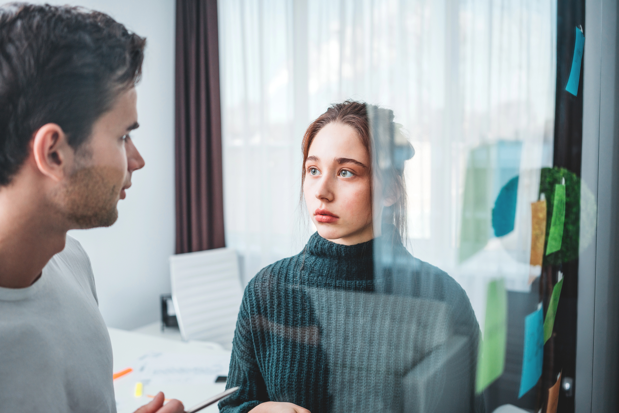 A man and a woman are having a conversation near a window in an office setting. The woman, wearing a dark sweater, looks attentively at the man. Sticky notes are visible on the glass. The room is softly lit by natural light.