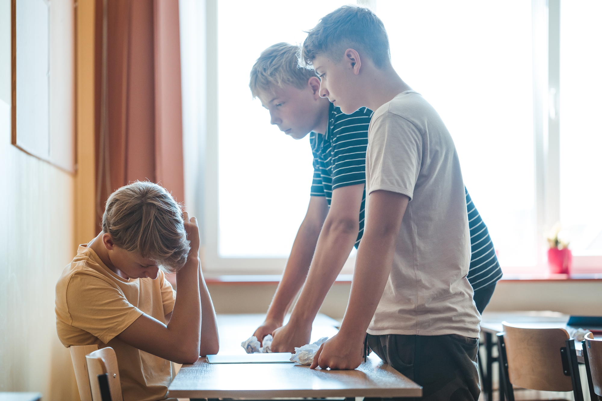 Three boys are in a classroom with sunlight streaming through the window. Two boys are standing over a seated boy who appears upset, resting his head on his hand. The atmosphere suggests a tense or difficult interaction.