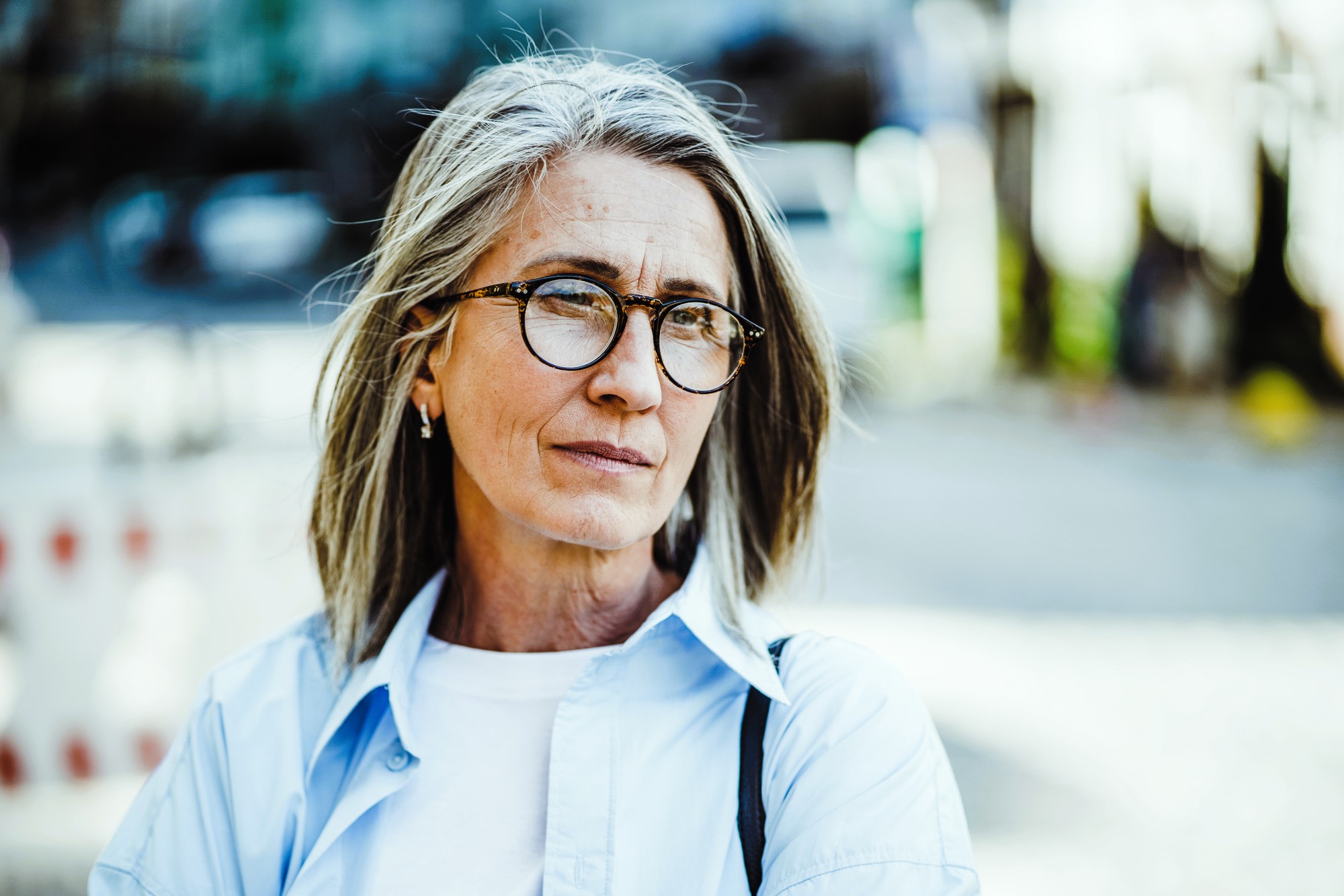 A woman with long gray hair and glasses stands outdoors, wearing a white shirt and light blue jacket. She looks thoughtful, with a blurred background of city elements in natural light.