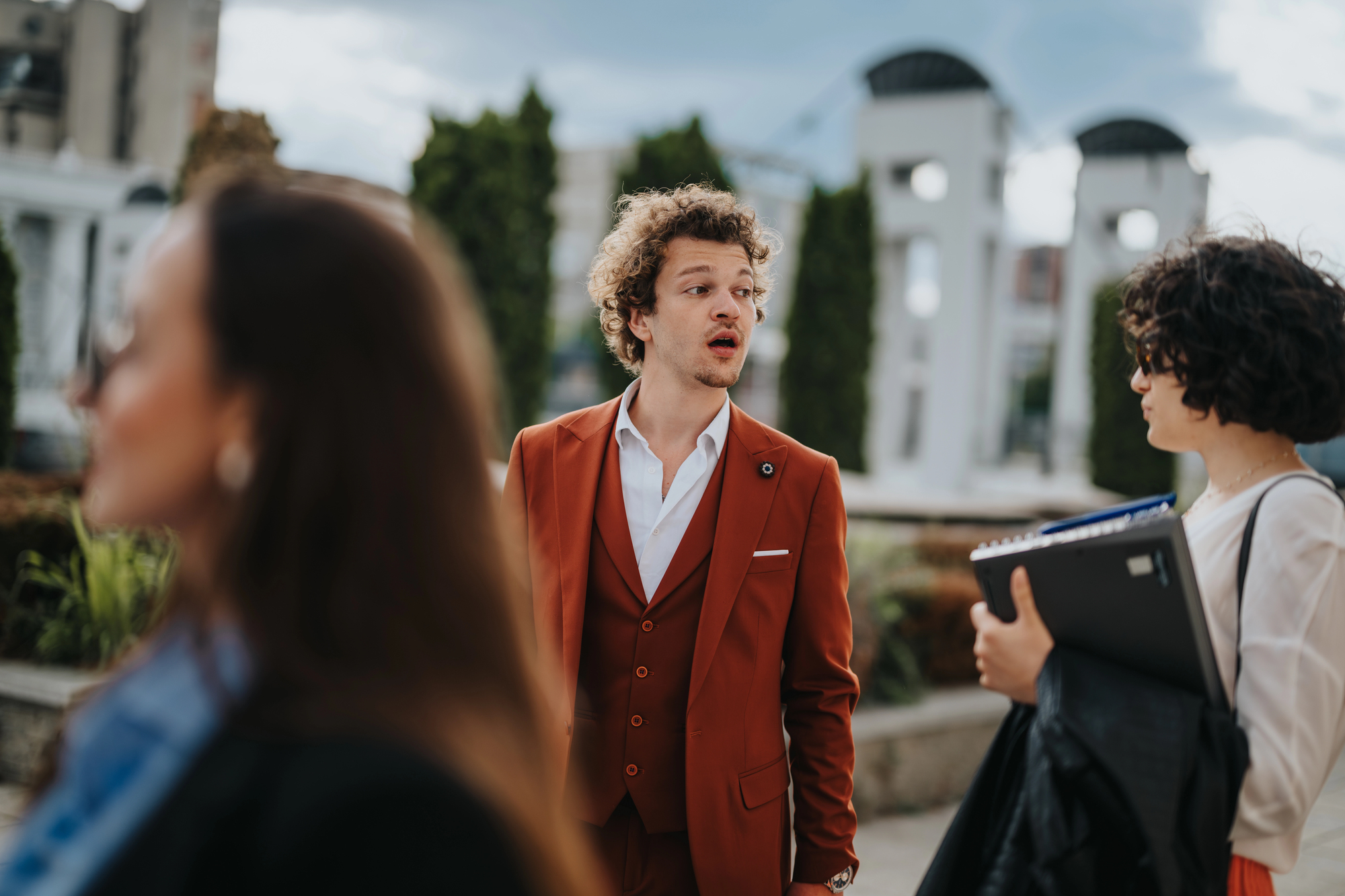 A man in a red suit stands outdoors, looking to the side with a surprised expression. He is surrounded by two people, one blurred in the foreground and another holding a folder. Background shows trees and modern structures.