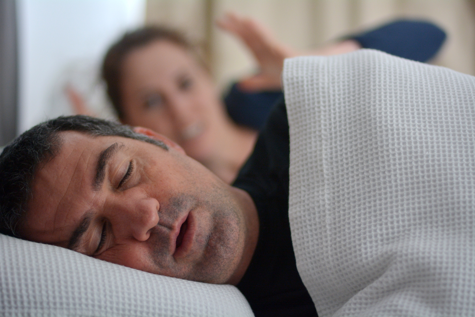 A man is sleeping on his side with his mouth slightly open, covered by a white blanket. In the background, a woman is lying on her back with her legs up, looking towards him. The setting appears to be a bedroom.