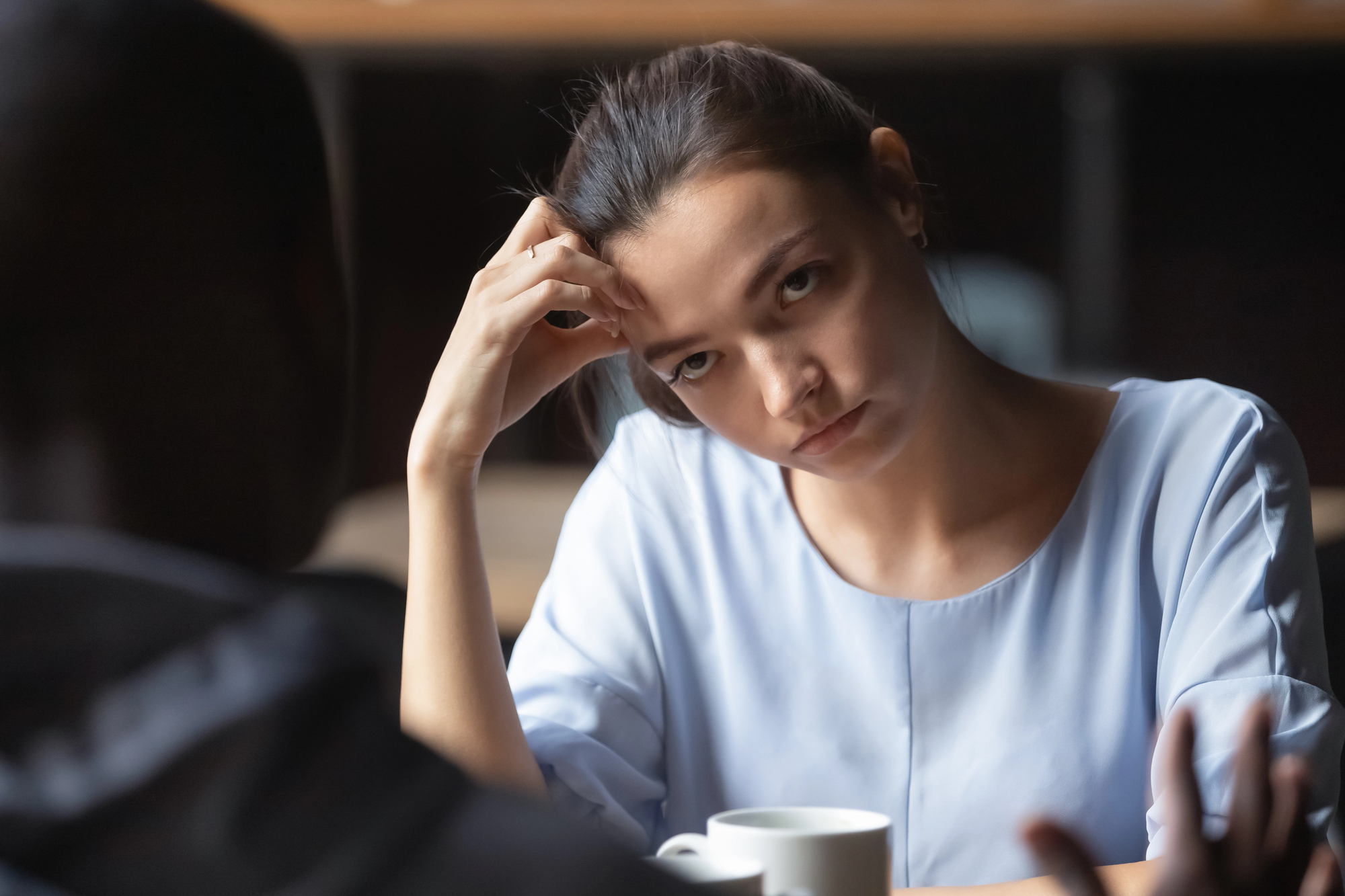 A woman wearing a light blue top sits at a table with her hand resting on her forehead, looking intently at someone across from her. A cup is in front of her, and the background is softly blurred.