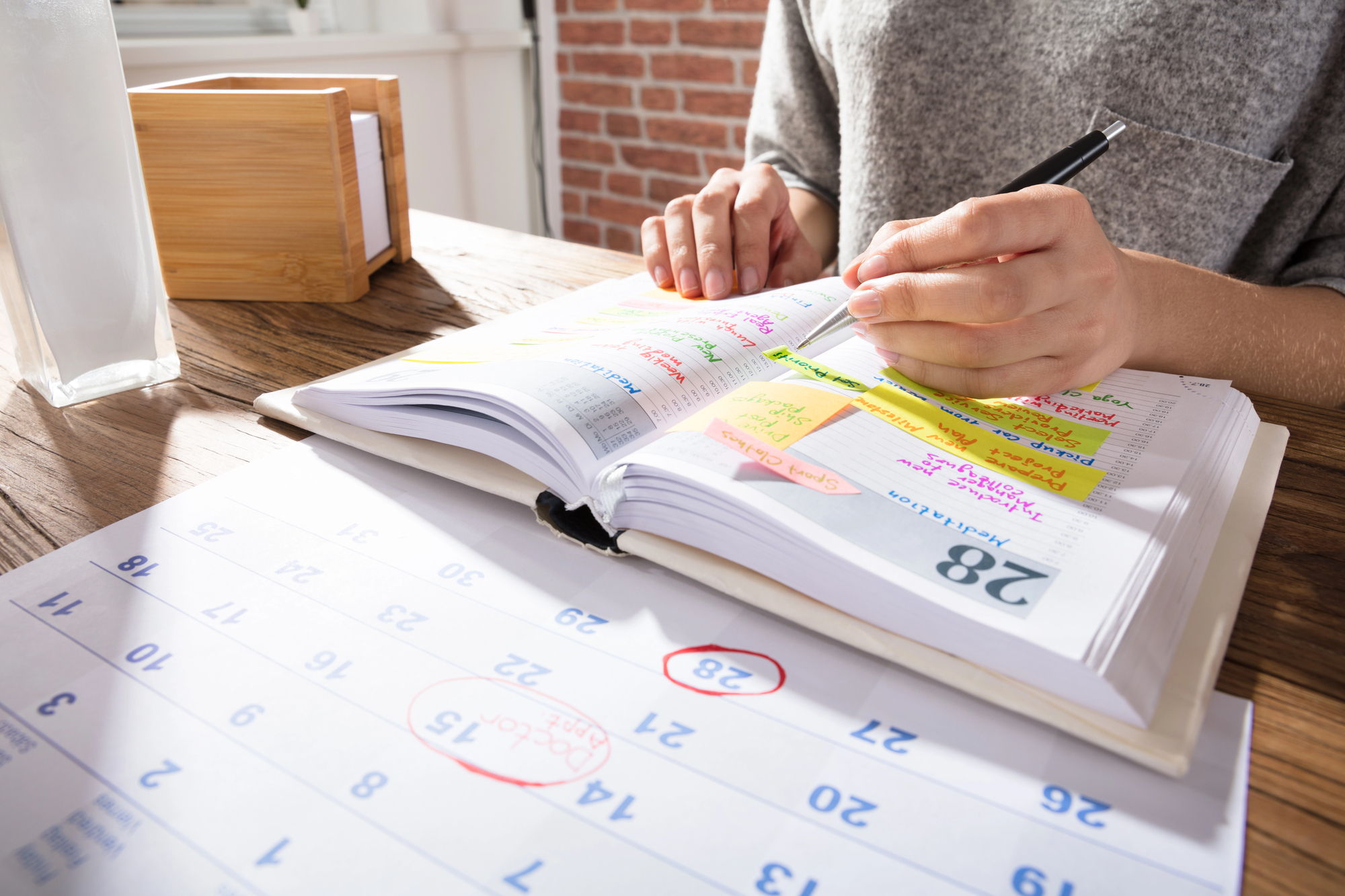 A person writing in a planner with colorful sticky notes and a pen. A calendar page with circled dates is on the wooden table. Sunlight and a small wooden box are in the background, with a brick wall partially visible.