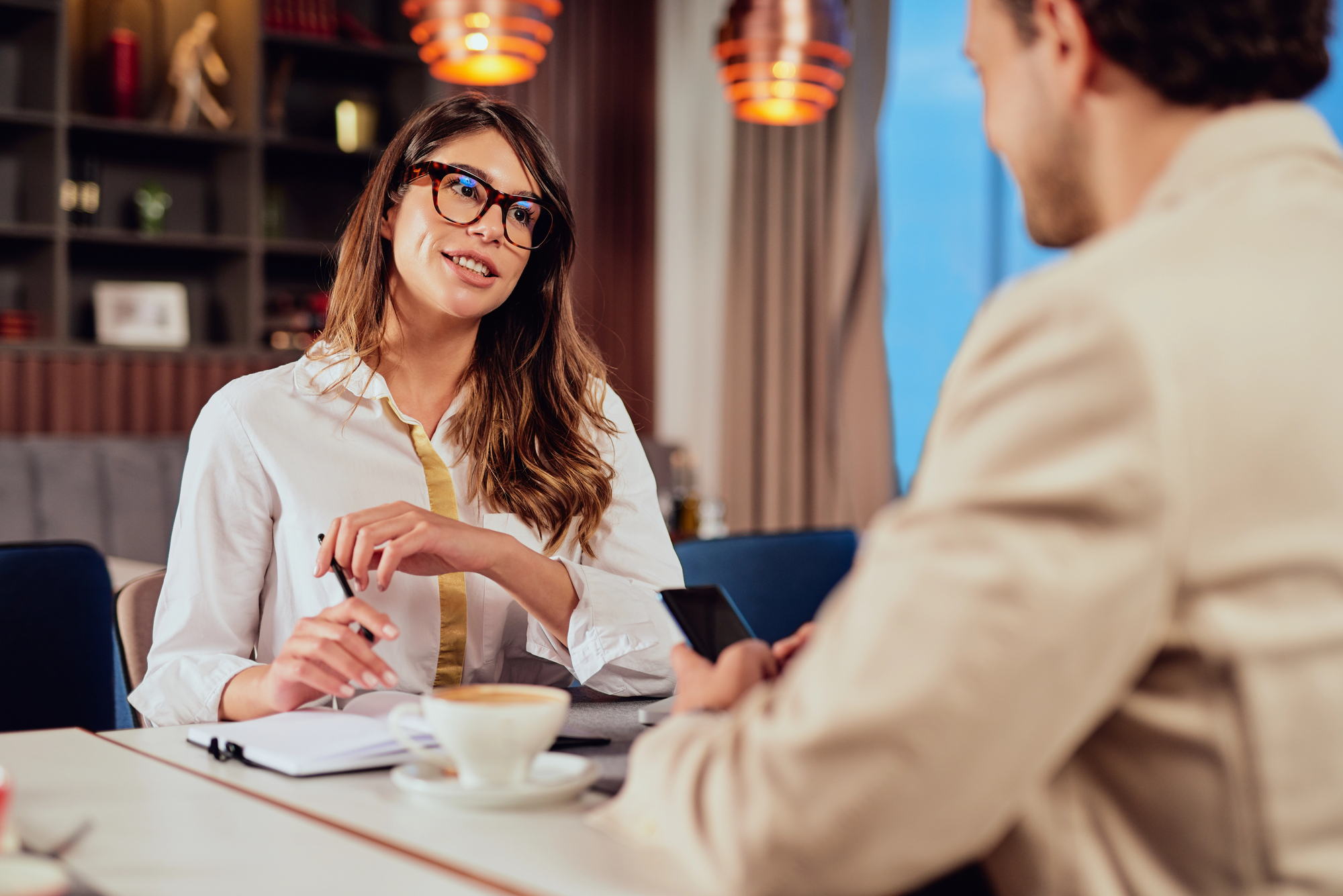 A woman in glasses is smiling and holding a phone during a conversation with a man. They are seated at a table with a coffee cup. The setting appears to be a cozy restaurant or cafe with warm lighting and shelves in the background.