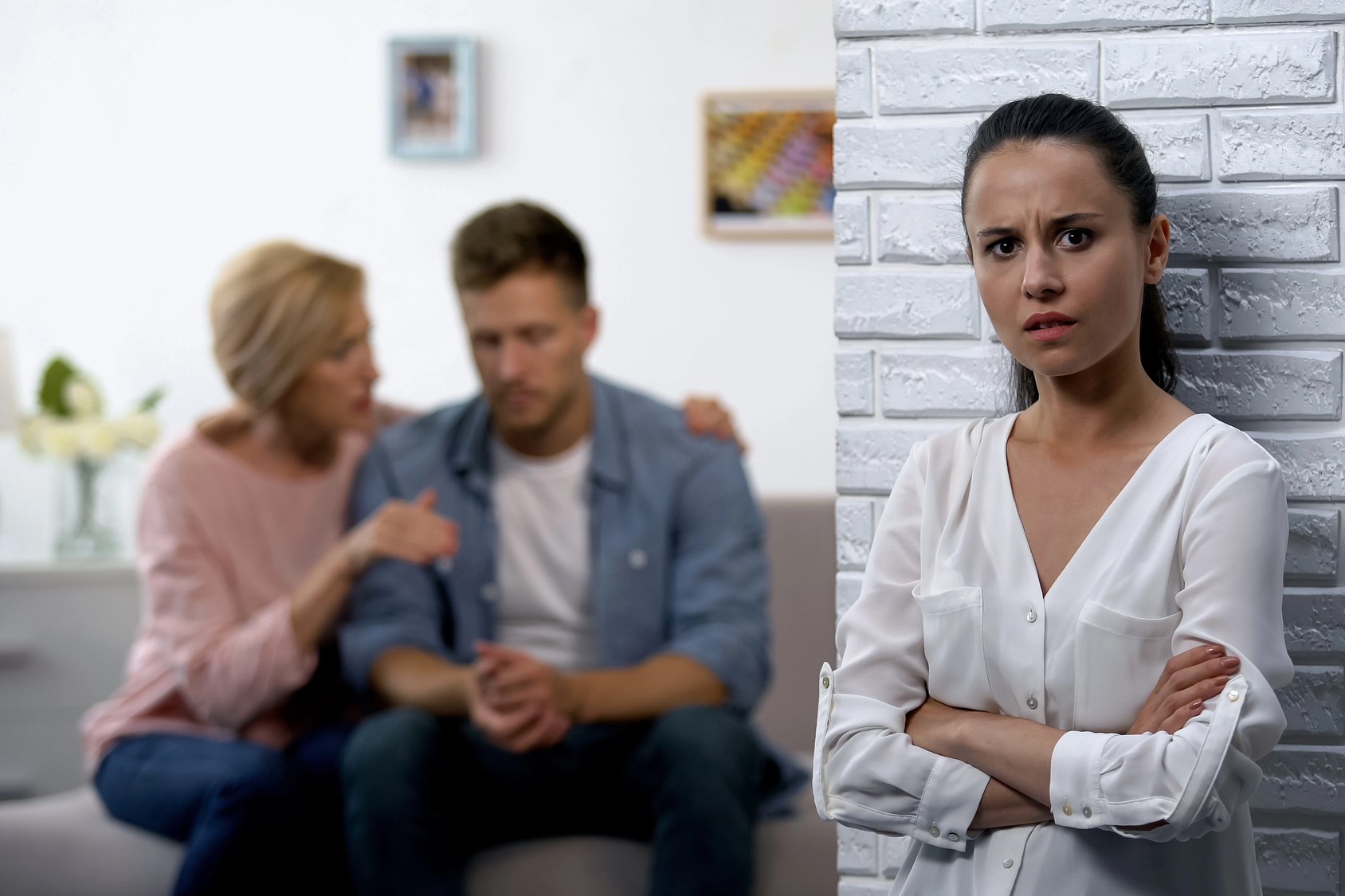 A woman stands with arms crossed, looking concerned. In the blurred background, a man sits on a couch, head down, while an older woman sits beside him, gesturing as if consoling. The setting is a cozy room with decorative elements.