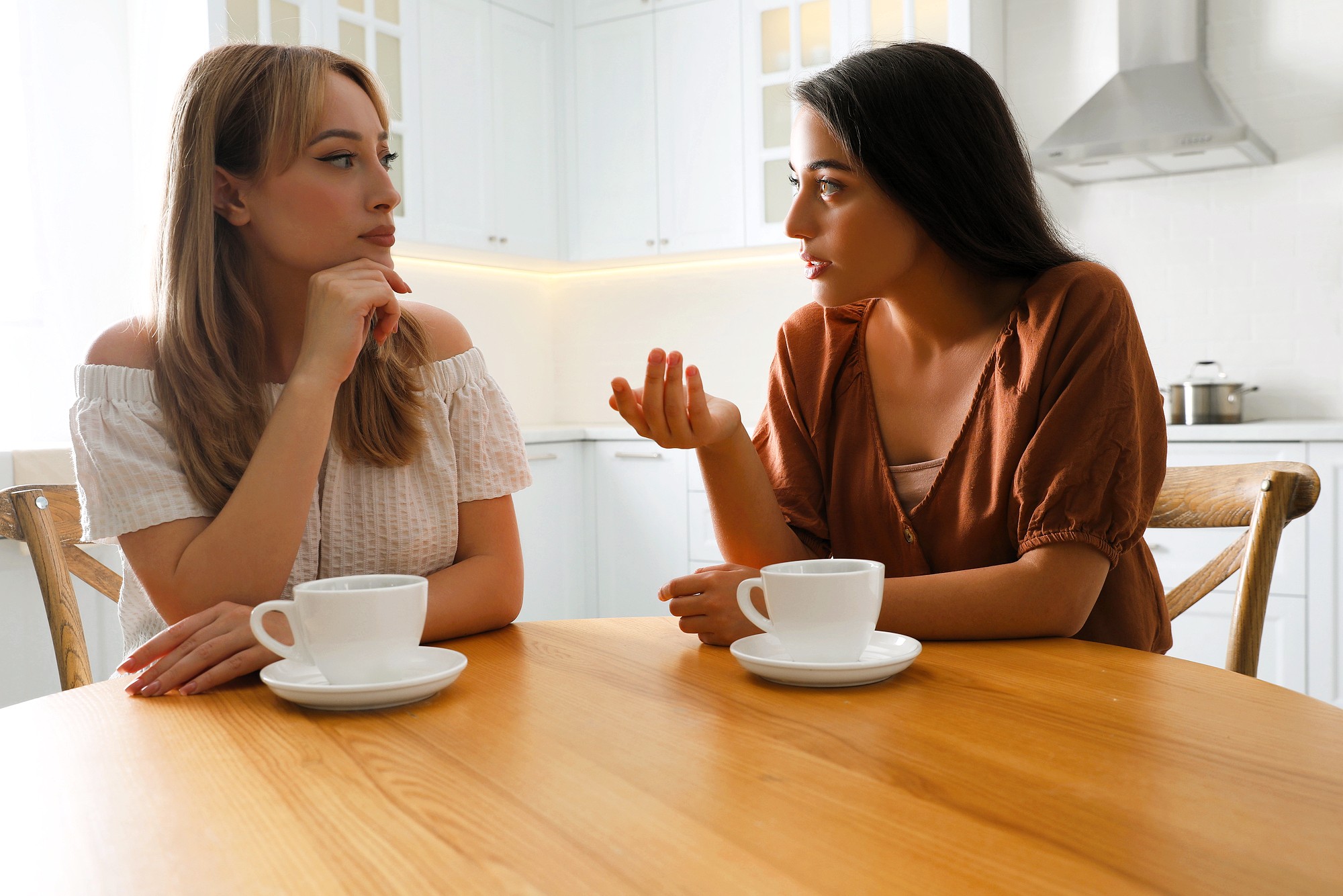 Two women sit at a wooden table in a bright kitchen, engaged in conversation. One gestures with her hand while speaking, and both have cups of coffee in front of them. The kitchen features white cabinets and a stovetop in the background.