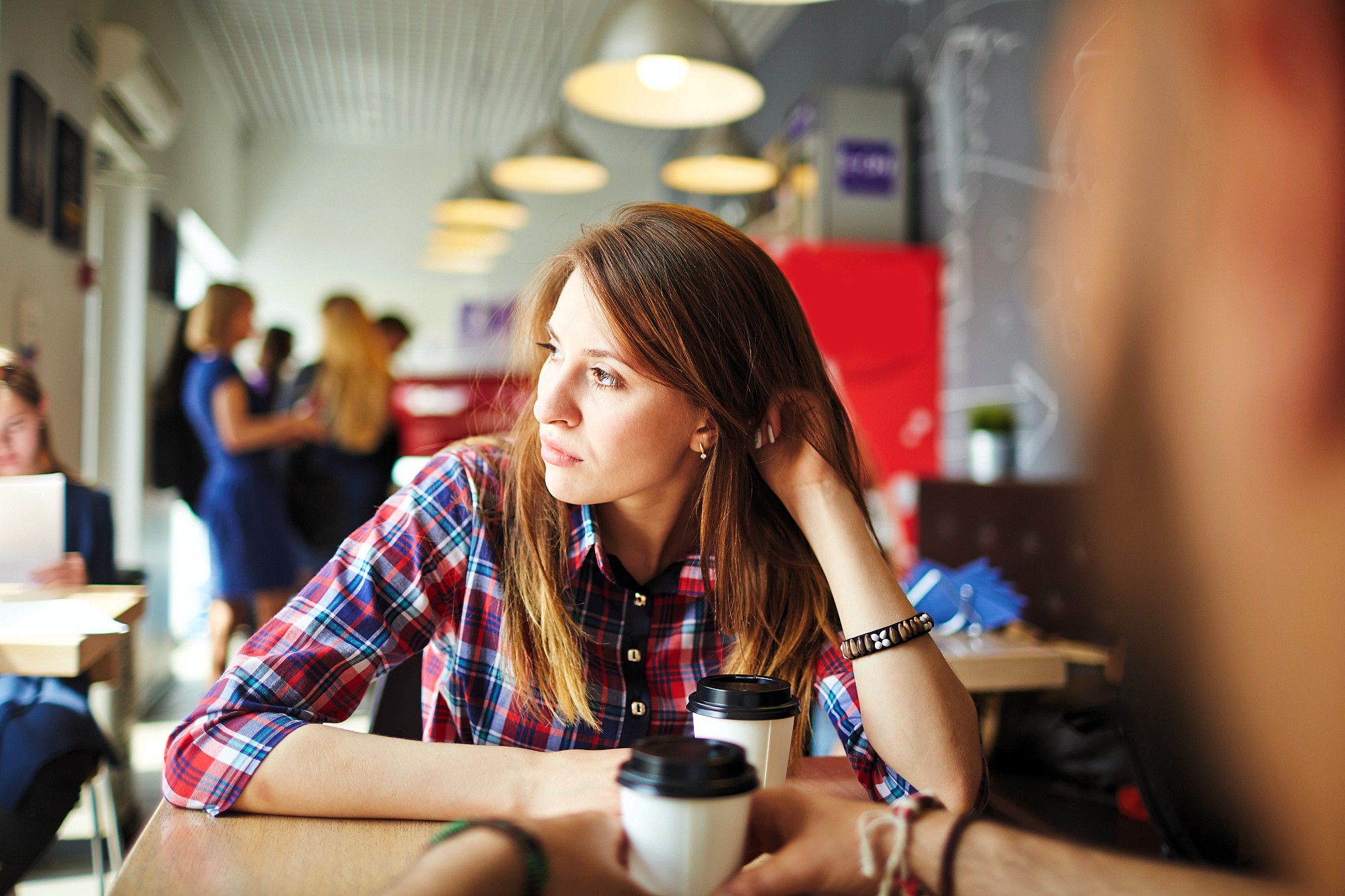 A woman in a plaid shirt sits at a table in a cafe, looking out a window with a thoughtful expression. She holds a cup of coffee. In the blurry background, people are engaged in conversation and activity.