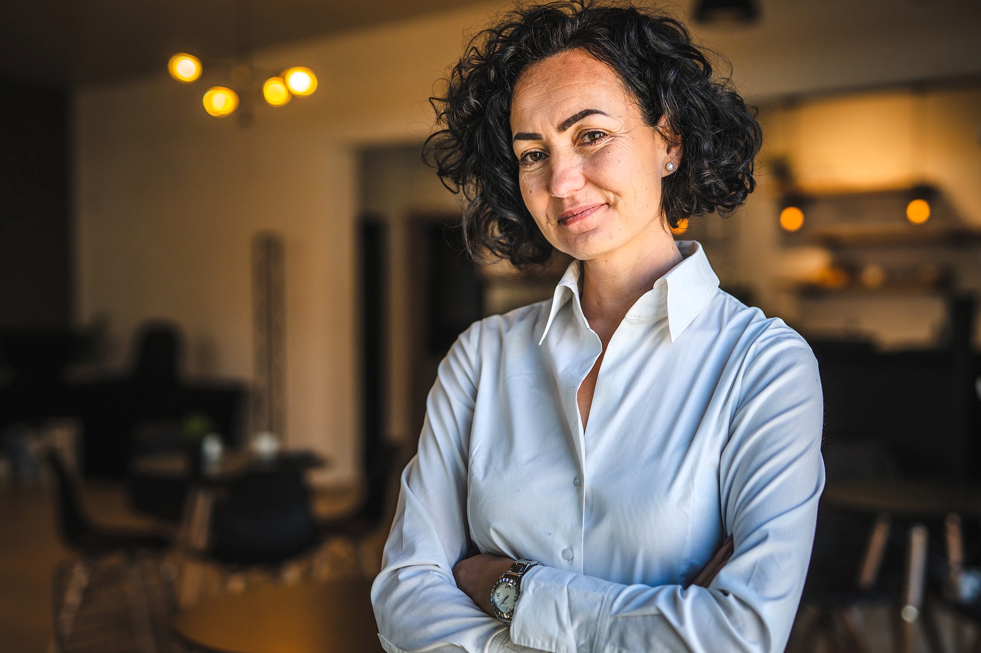 Woman with curly hair and a white blouse smiles confidently while standing with arms crossed in a warmly lit room. The background includes blurred furnishings and soft lighting.