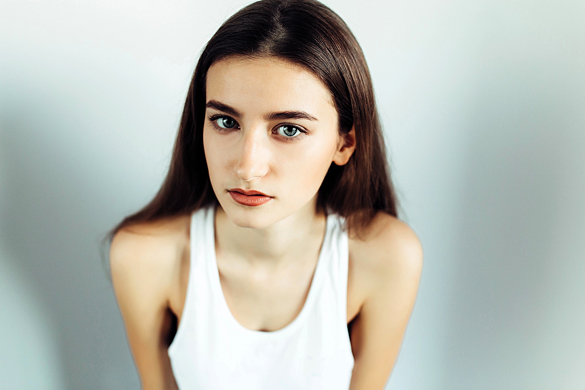 A woman with long brown hair and green eyes is looking directly at the camera. She is wearing a sleeveless white top and is set against a plain background. The lighting highlights her facial features and expression.