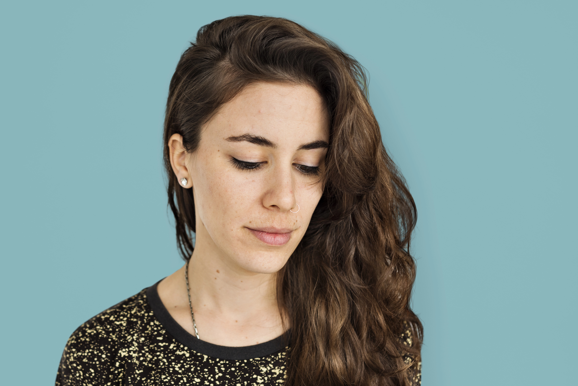 Woman with long, wavy brown hair looks down against a solid light blue background. She is wearing a black top with golden speckles and small stud earrings. Her expression is calm and contemplative.