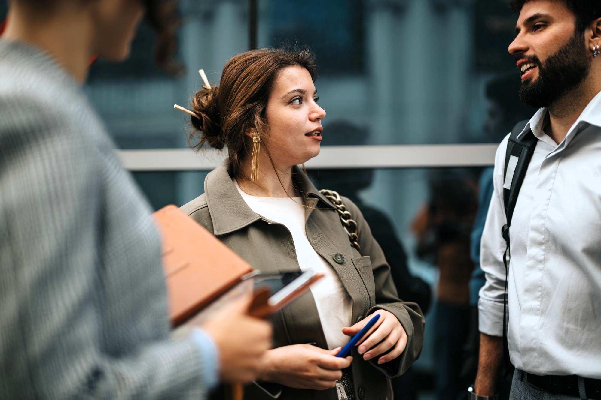 Three people are standing and talking near a glass wall. The woman in the center is holding a pen, while another person in the foreground holds a folder and phone. A man with a beard stands on the right, smiling.