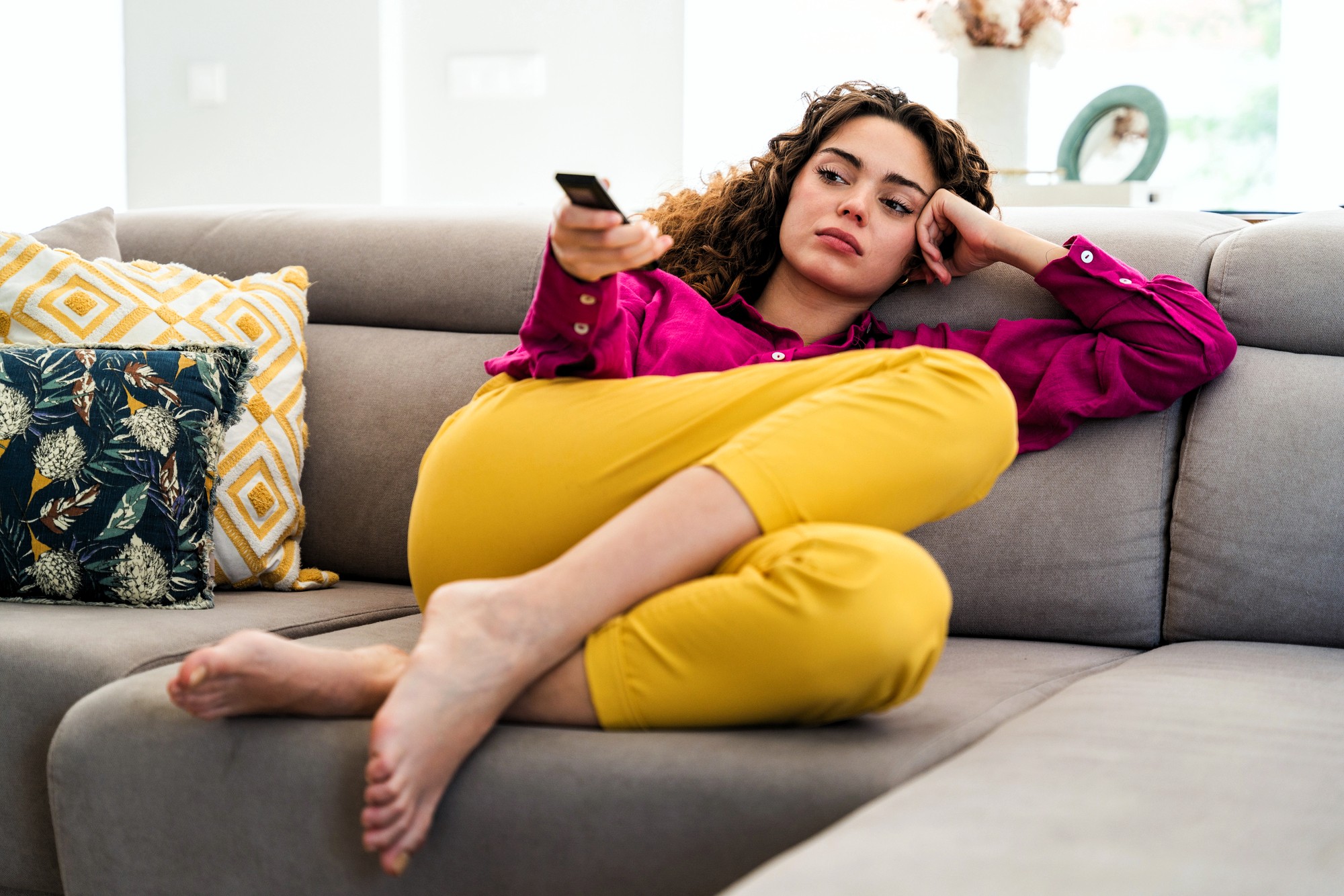A woman in a pink shirt and yellow pants lounges on a sofa, holding a TV remote and looking bored or pensive. Patterned cushions are beside her, and she is barefoot with legs curled up.