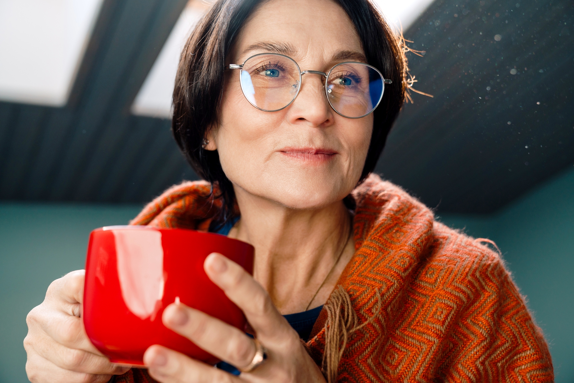 A person with short dark hair and glasses is holding a red mug while wearing an orange patterned shawl. They are looking thoughtfully to the side, with light coming from a skylight above.
