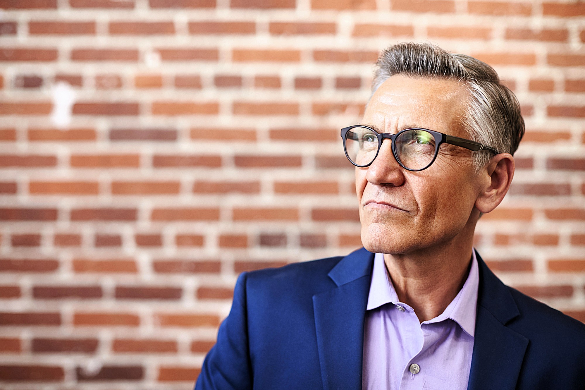 A mature man wearing glasses and a navy blazer over a lavender shirt stands against a brick wall, looking thoughtfully into the distance.