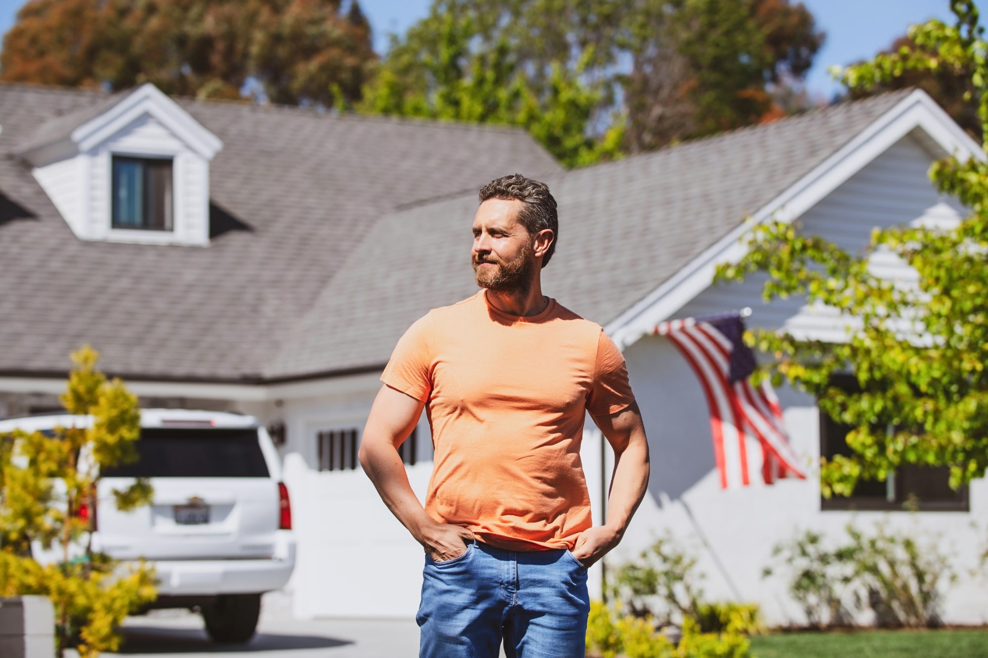 A man wearing an orange t-shirt and blue jeans stands outside with his hands in his pockets, in front of a white house with an American flag and a white SUV parked in the driveway. Green trees surround the sunny scene.