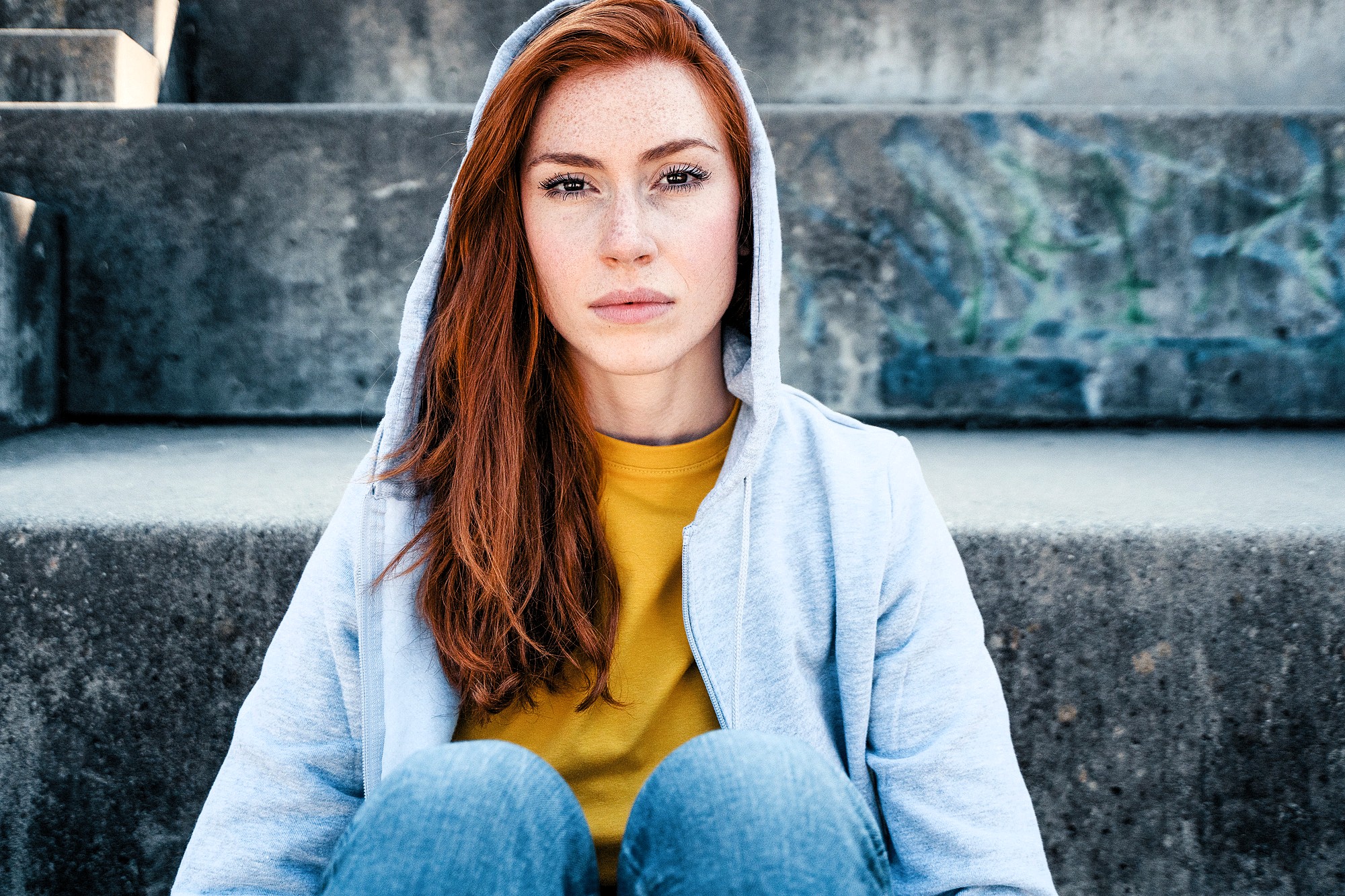 A young woman with long red hair, wearing a light gray hoodie and yellow shirt, sits on concrete steps outdoors, looking directly at the camera with a neutral expression.