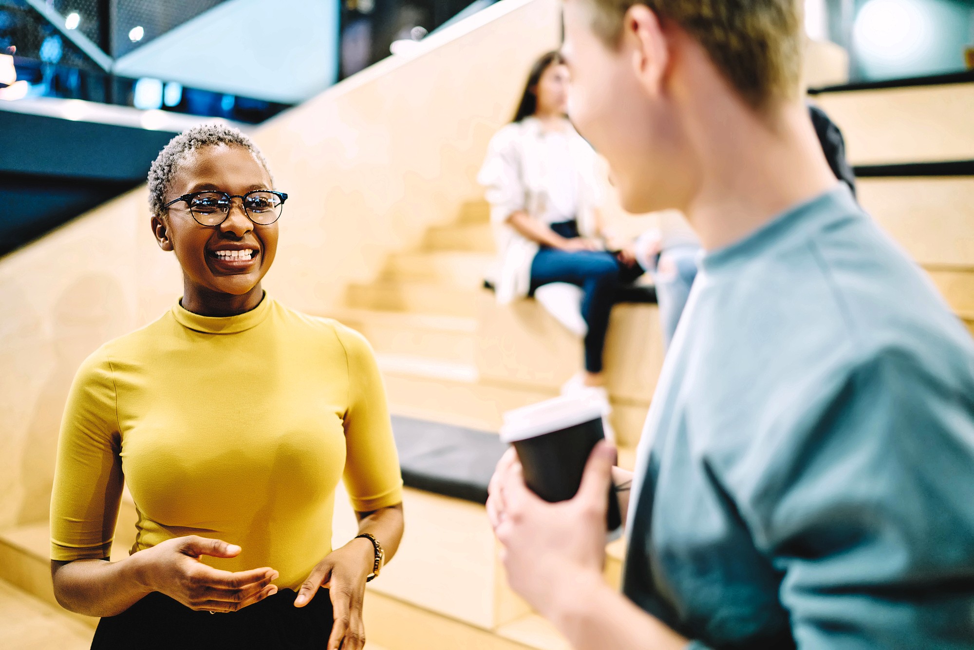 A woman in a yellow top and glasses smiles while talking to a man holding a coffee cup. They are standing in a modern, indoor setting with wooden steps and people in the background.
