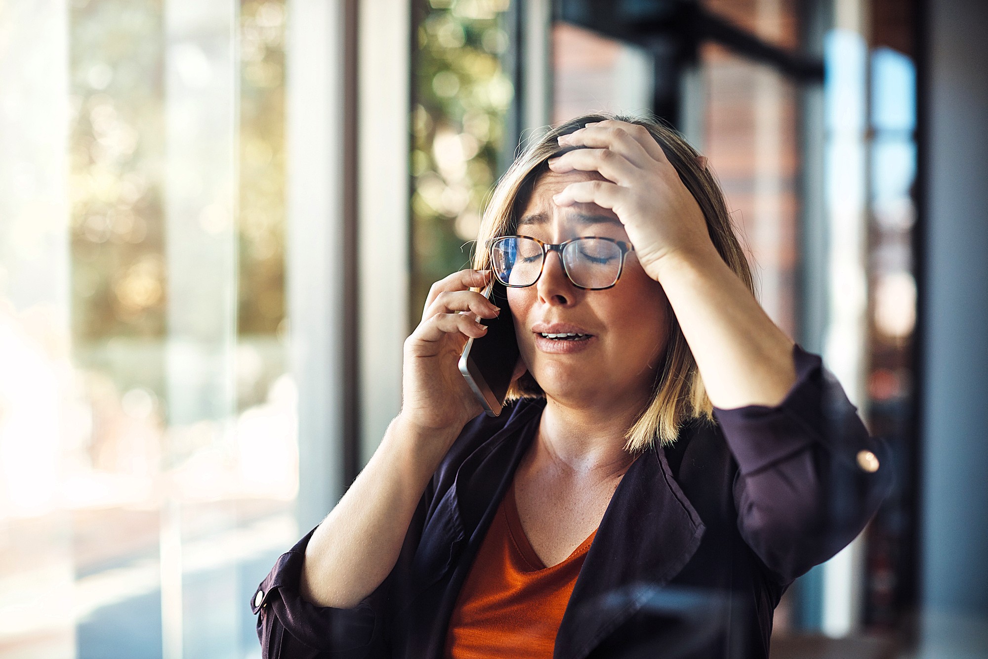 A woman with short blonde hair and glasses is standing by a window, holding a phone to her ear with one hand and touching her forehead with the other. She appears distressed, with her eyes closed. Sunlight streams in through the window behind her.