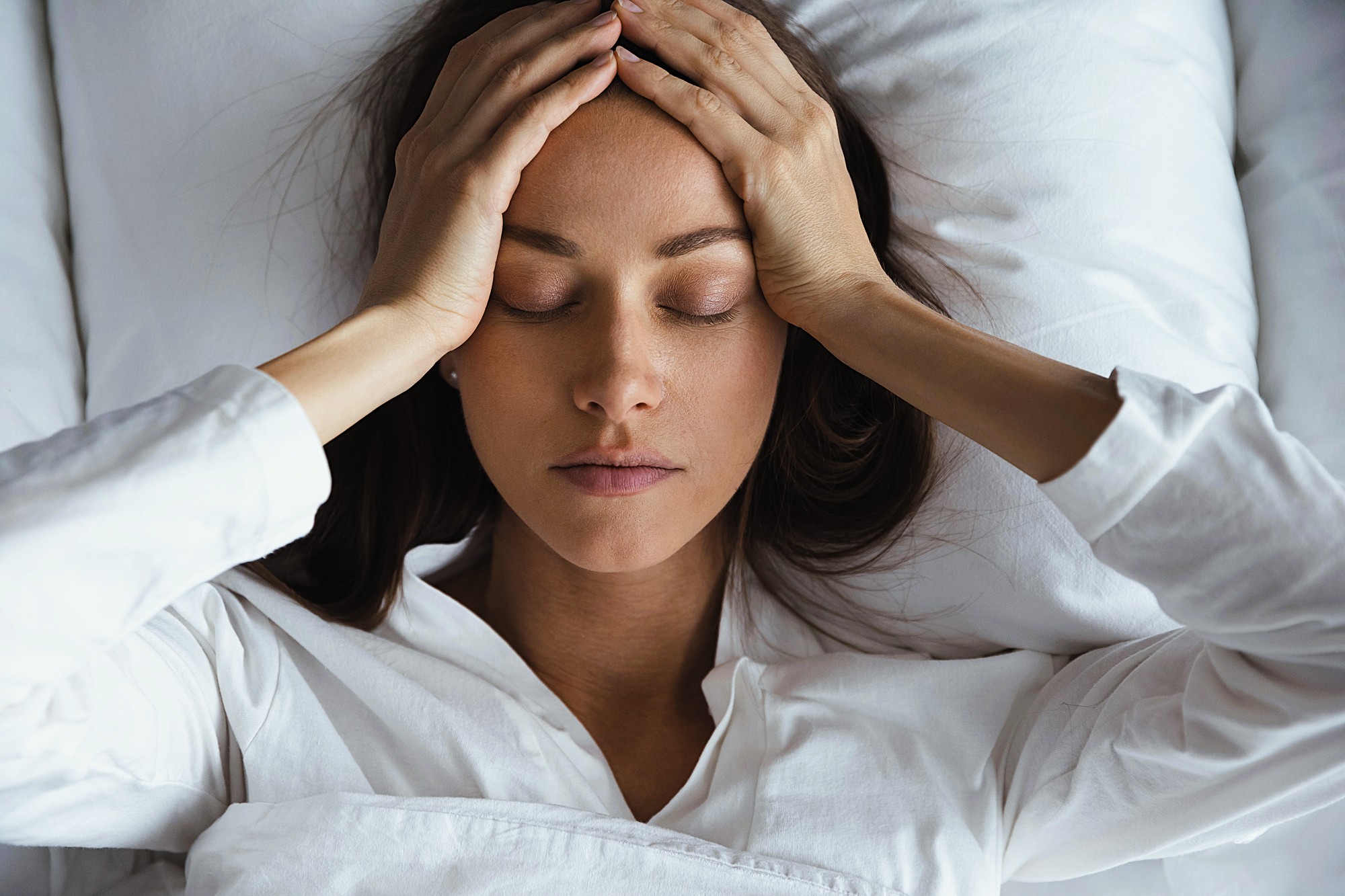 A woman lies in bed with her eyes closed, holding her hands to her forehead, appearing stressed or unwell. She is wearing a white long-sleeve shirt and is partially covered by a white blanket.