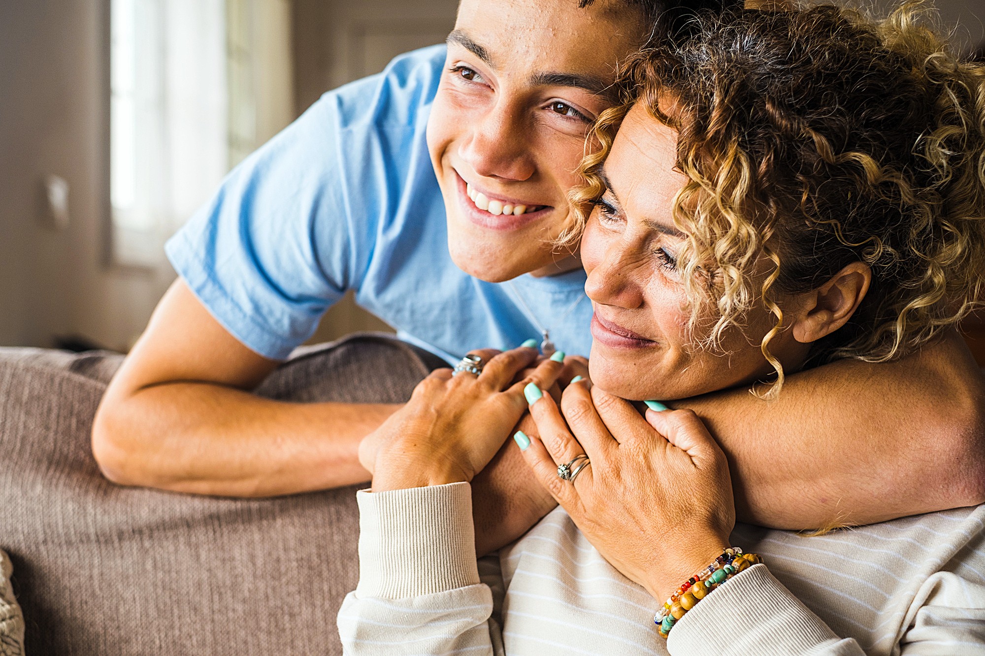 A young person in a blue shirt hugs a smiling adult from behind on a couch. The adult has curly hair and wears a light-colored sweater with colorful beaded bracelets. They both look happy and relaxed.