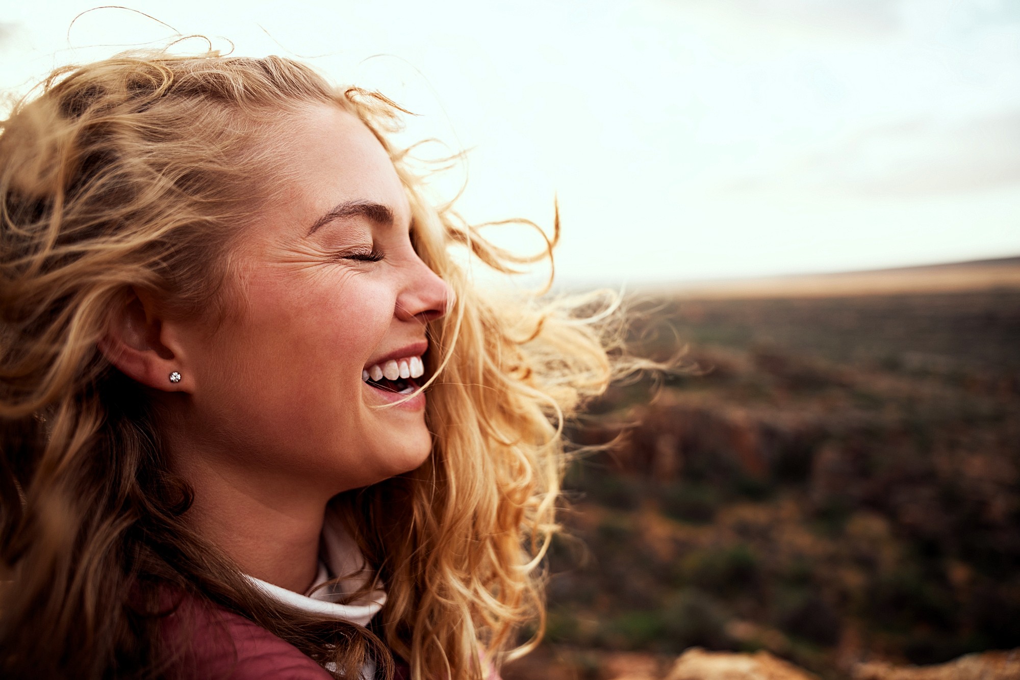 A young woman with long, wavy blonde hair smiles joyfully with her eyes closed outdoors. The wind blows her hair, and a blurred landscape stretches out behind her under a bright sky.