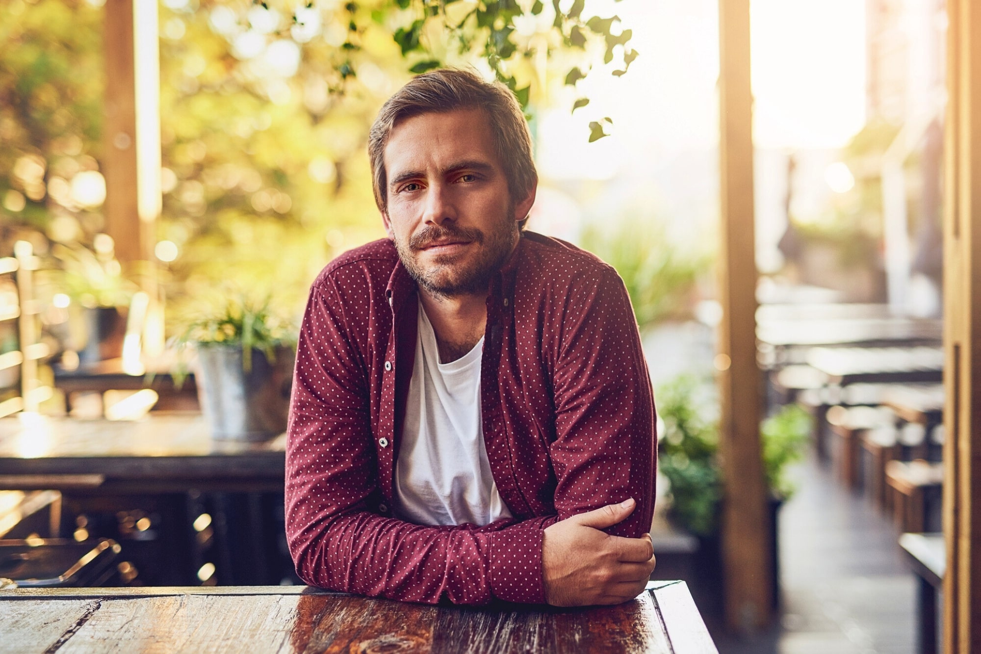 A man with short brown hair and a beard, wearing a maroon shirt over a white t-shirt, sits at a wooden outdoor table, smiling slightly. The background has plants and warm, natural light.
