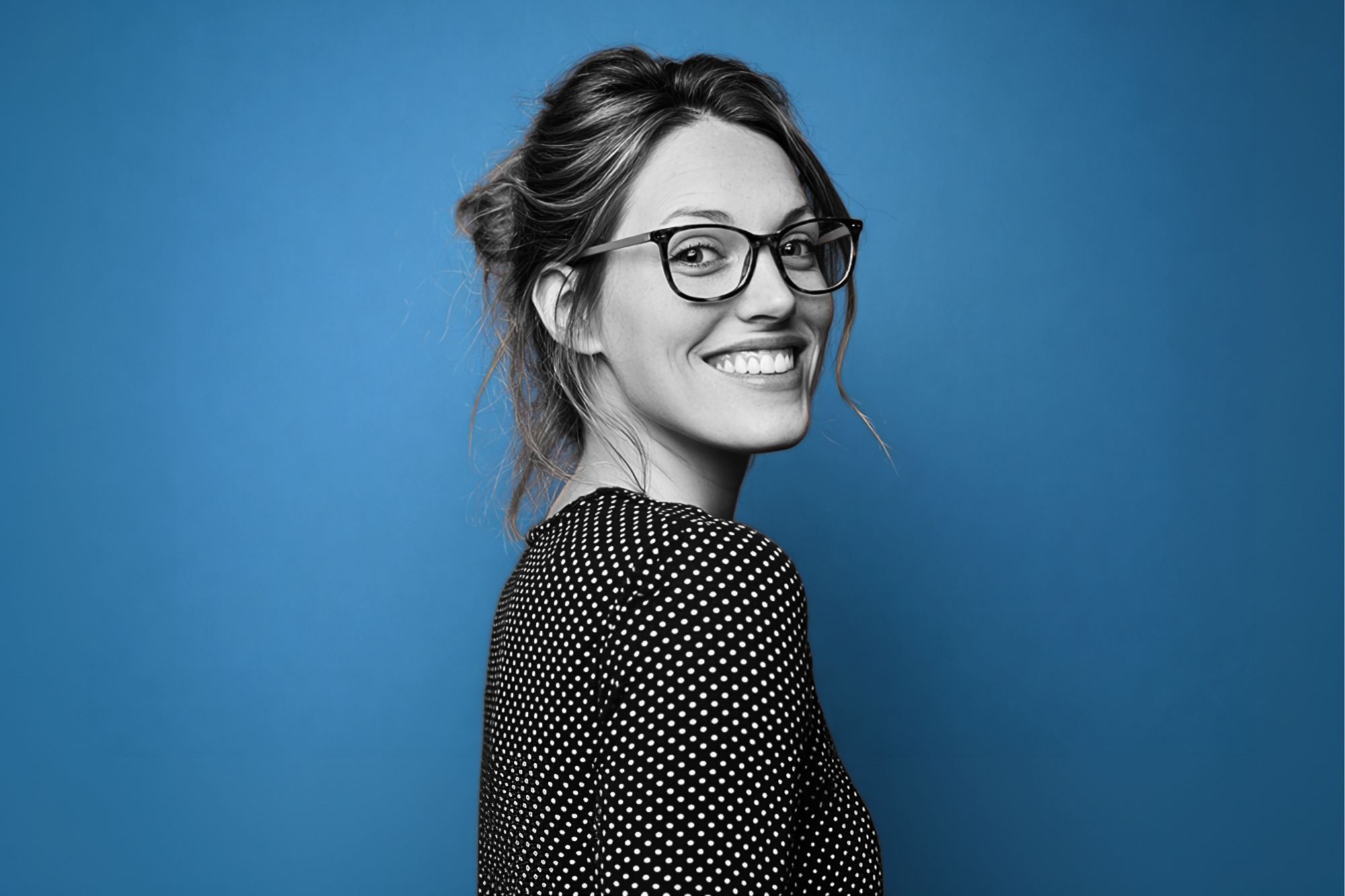 Woman with glasses and long hair tied back, smiling and looking over her shoulder. She is wearing a black and white polka dot top, with a blue background. The image is mostly black and white except for the background.