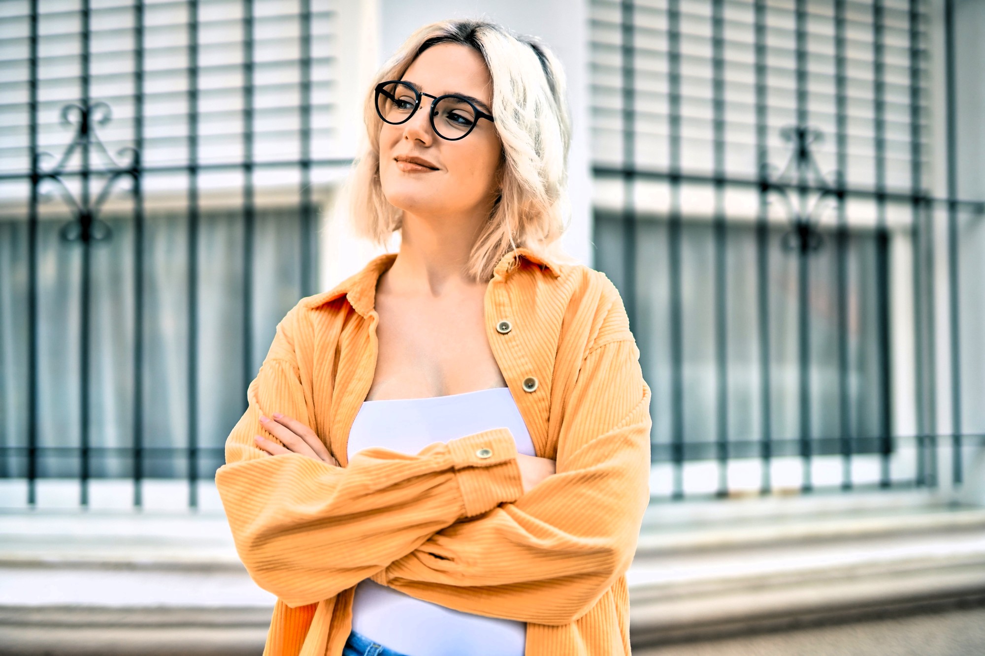 A person with short blond hair and glasses stands confidently, wearing an orange shirt over a white top. They are smiling slightly and have their arms crossed. The background features a window with metal bars.