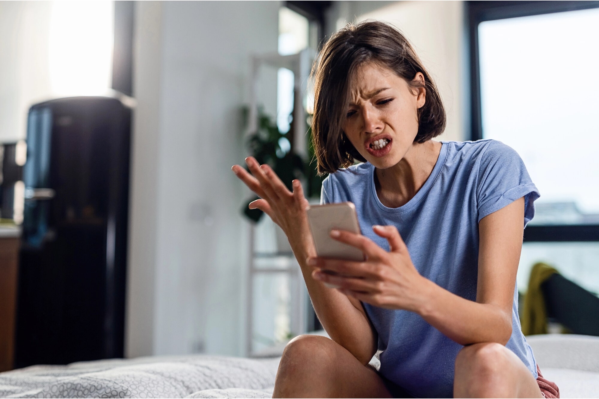 A young woman sitting indoors on a bed looks frustrated while holding a smartphone in one hand and gesturing with the other, her face showing irritation or anger.