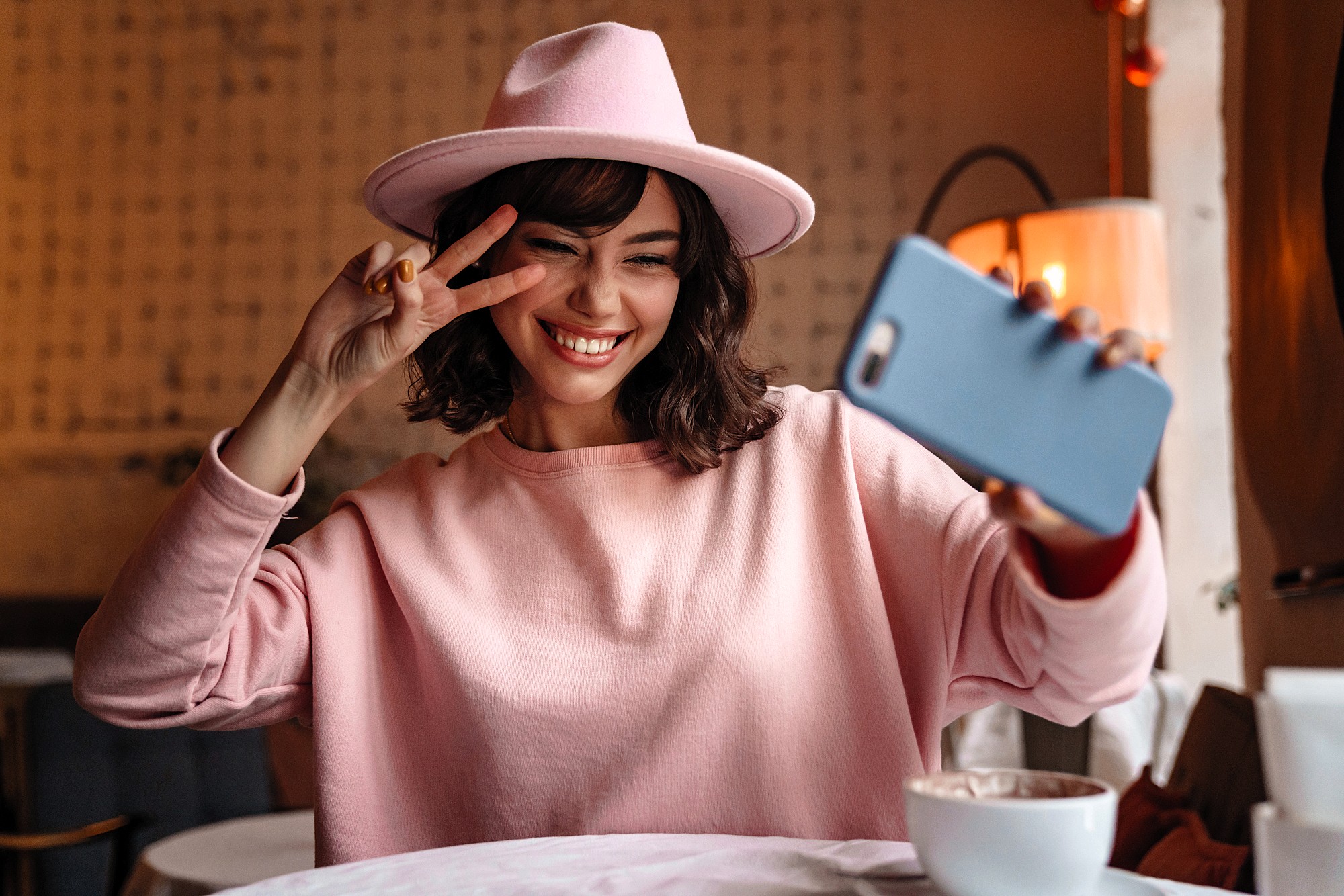 A smiling young woman in a pink sweater and hat takes a selfie with her phone, holding up a peace sign with her fingers, sitting at a table in a cozy café.