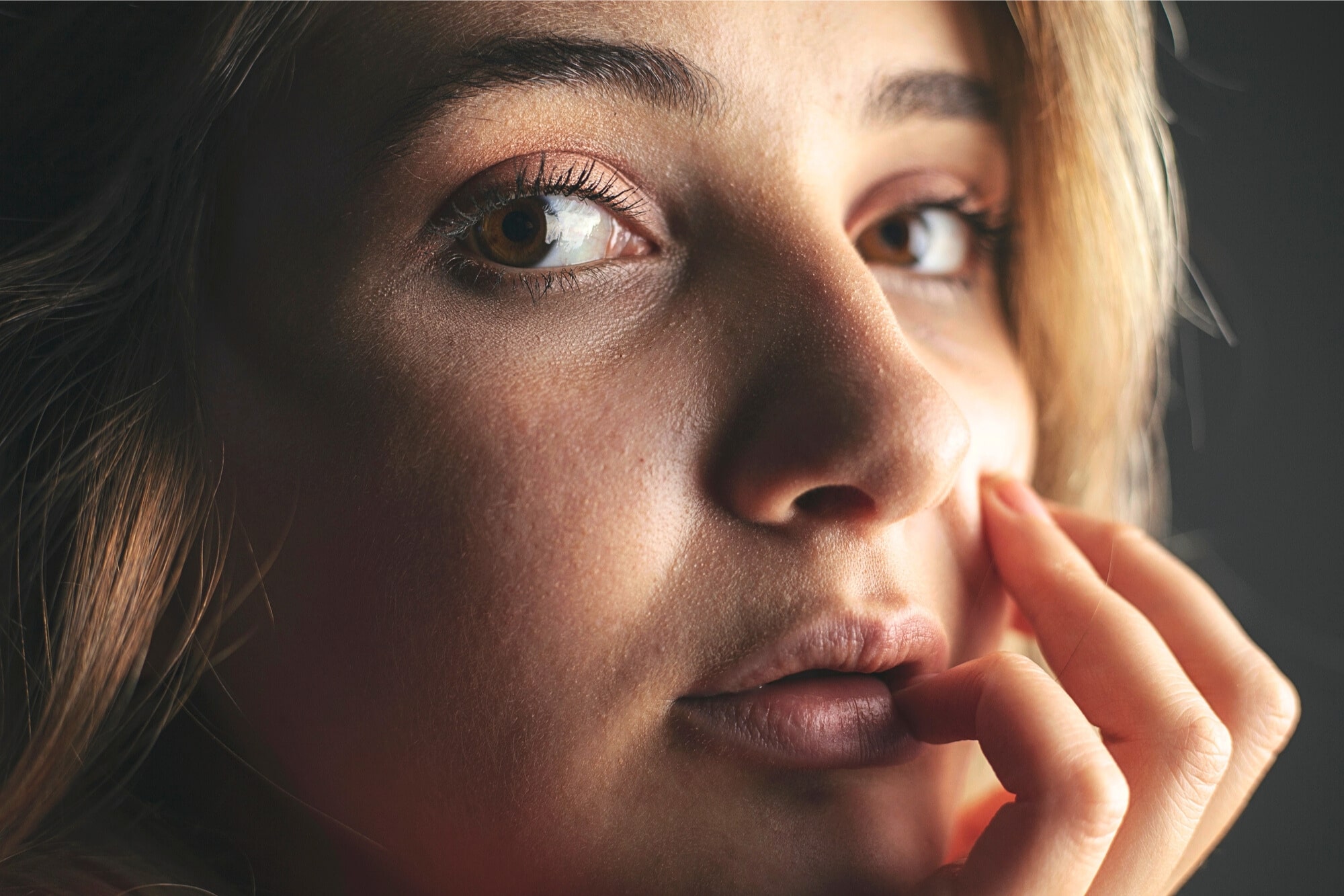 A close-up of a young person with light skin, light brown eyes, and blonde hair. They are looking thoughtfully at the camera, with their fingers gently touching their lips and cheek, in soft natural light.