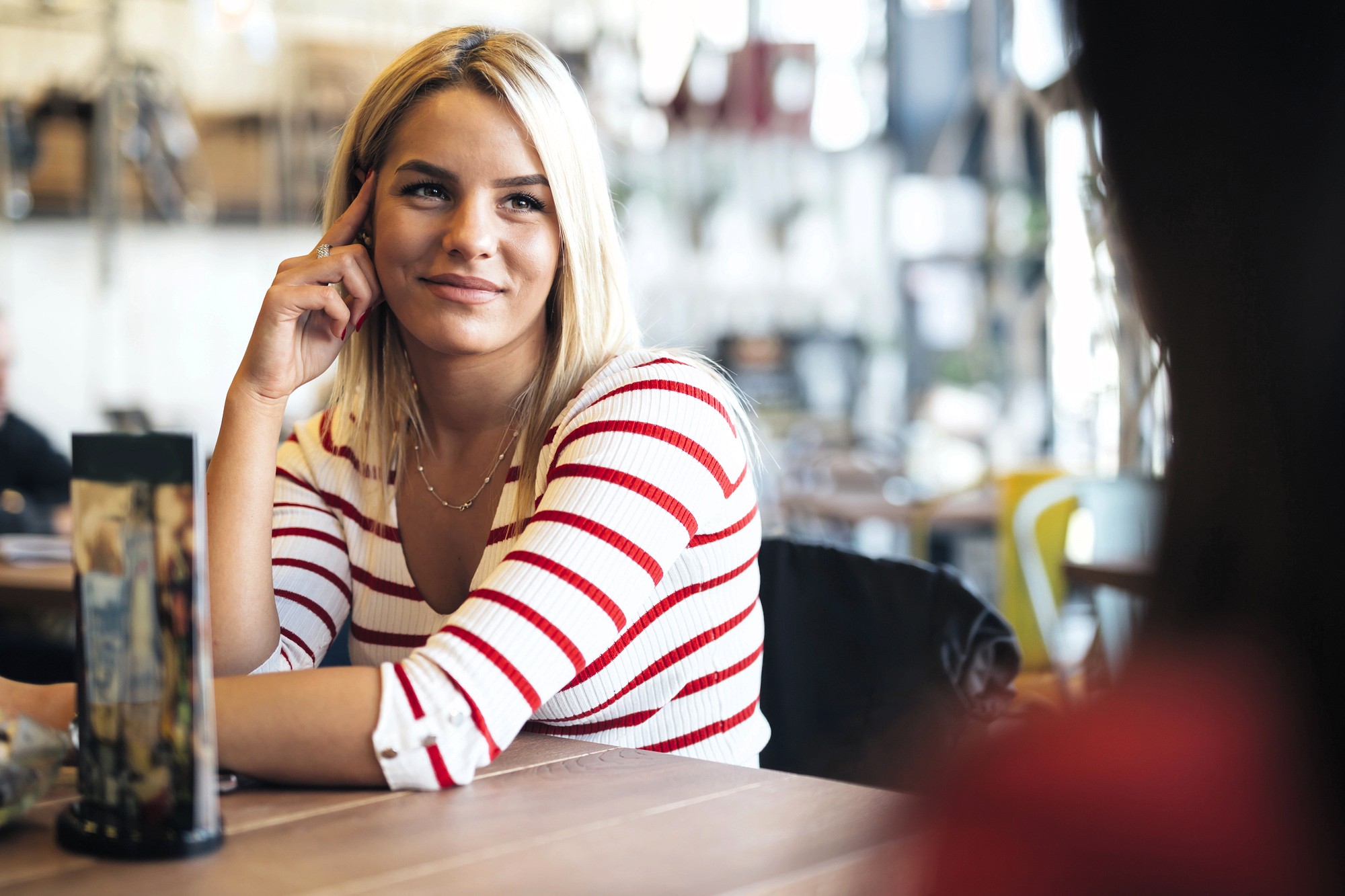 A woman with blonde hair wearing a white and red striped top sits at a table in a cafe, smiling and resting her head on her hand while listening to someone across from her.