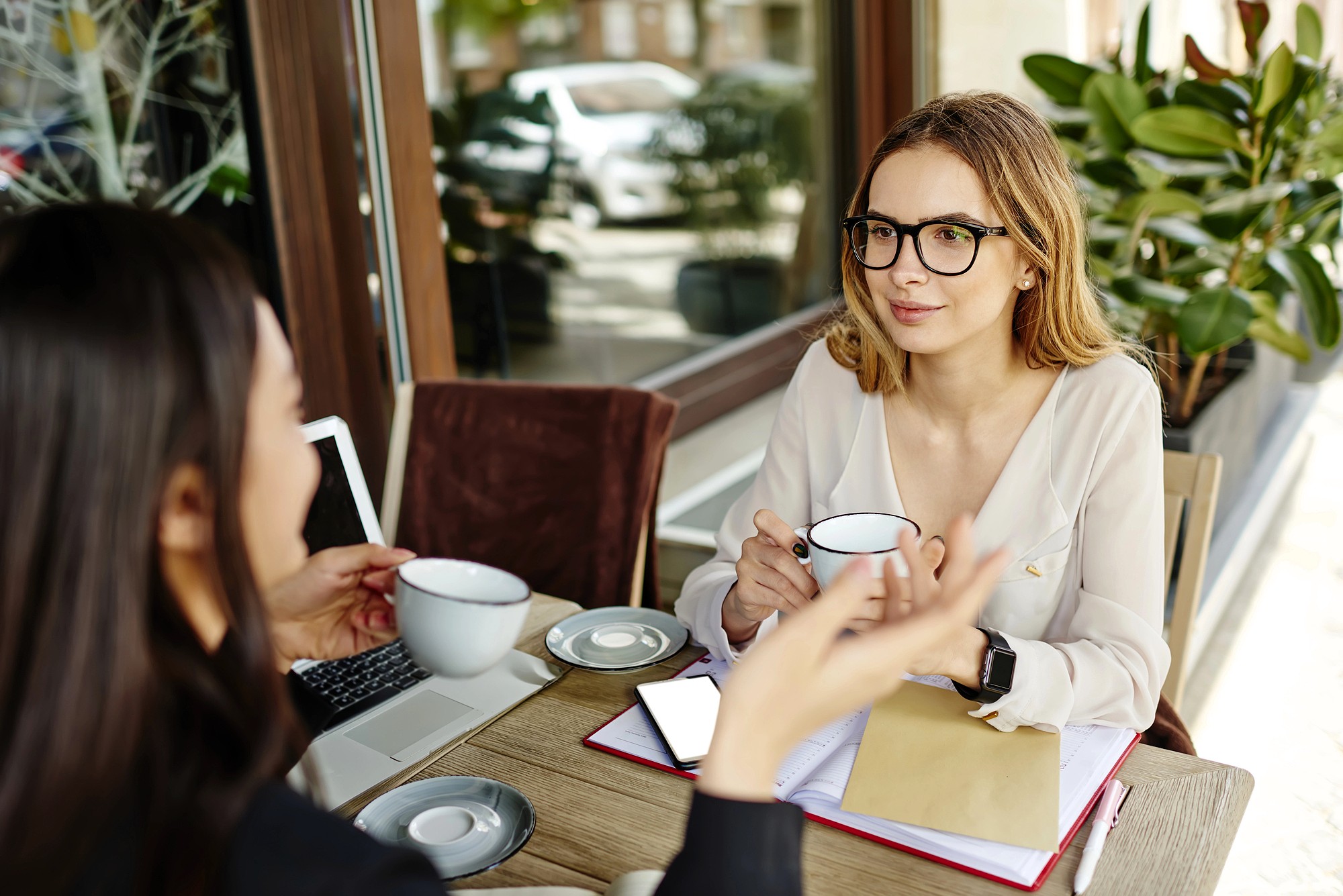 Two women sit at an outdoor café table, having a conversation over coffee. One woman faces the camera, wearing glasses, while the other is seen from behind. Notebooks, a laptop, and a phone are on the table.