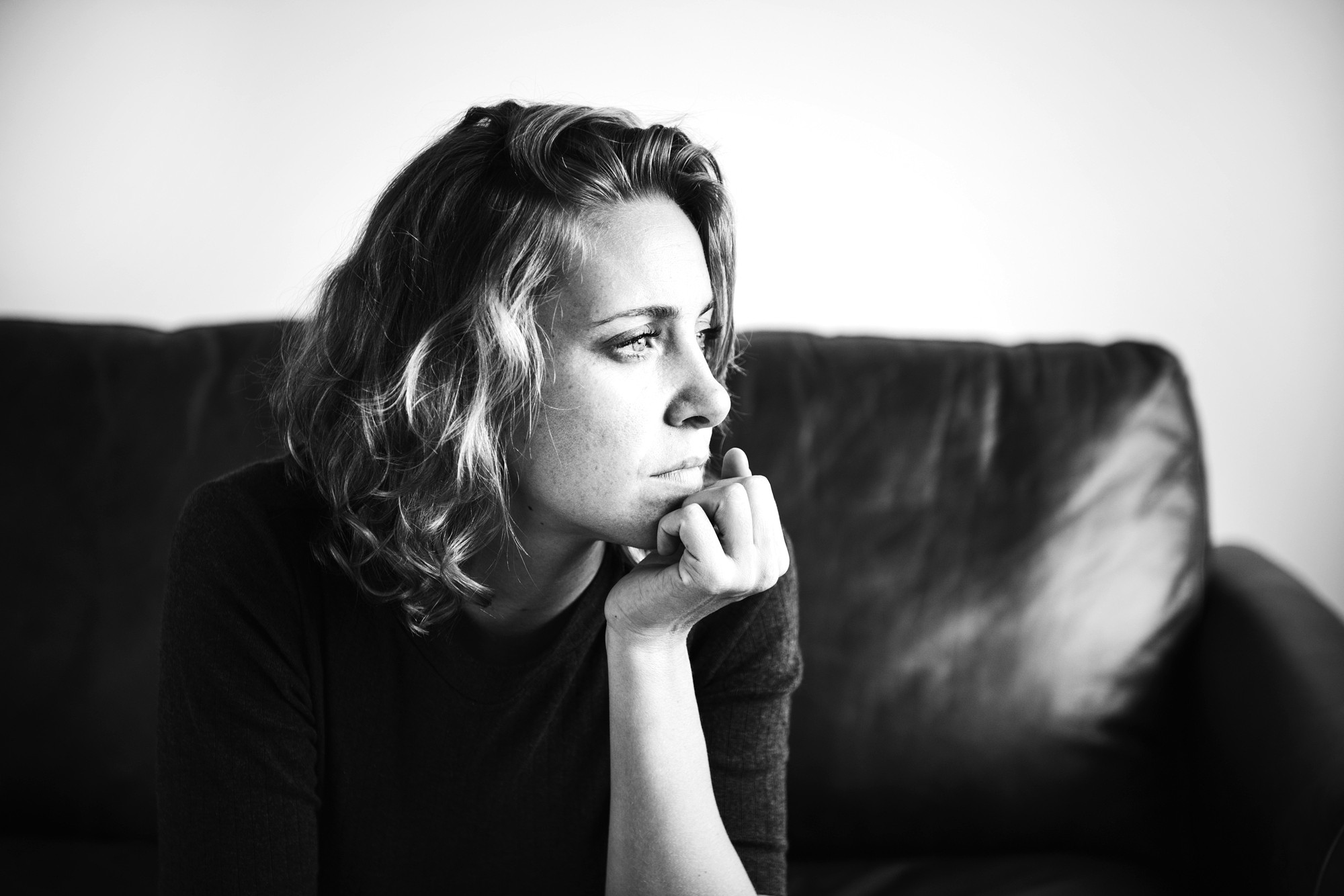 Black and white photo of a woman with short, wavy hair sitting on a couch, resting her chin on her hand, and gazing thoughtfully to the right. The background is plain and softly lit.