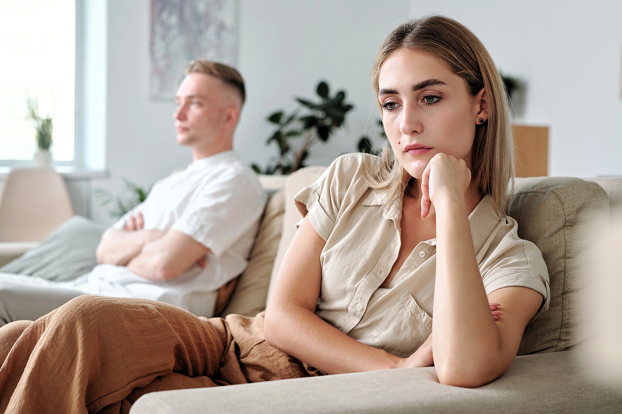 A woman sits on a couch looking sad and thoughtful, resting her chin on her hand, while a man sits in the background with his arms crossed, both appearing upset in a bright living room.