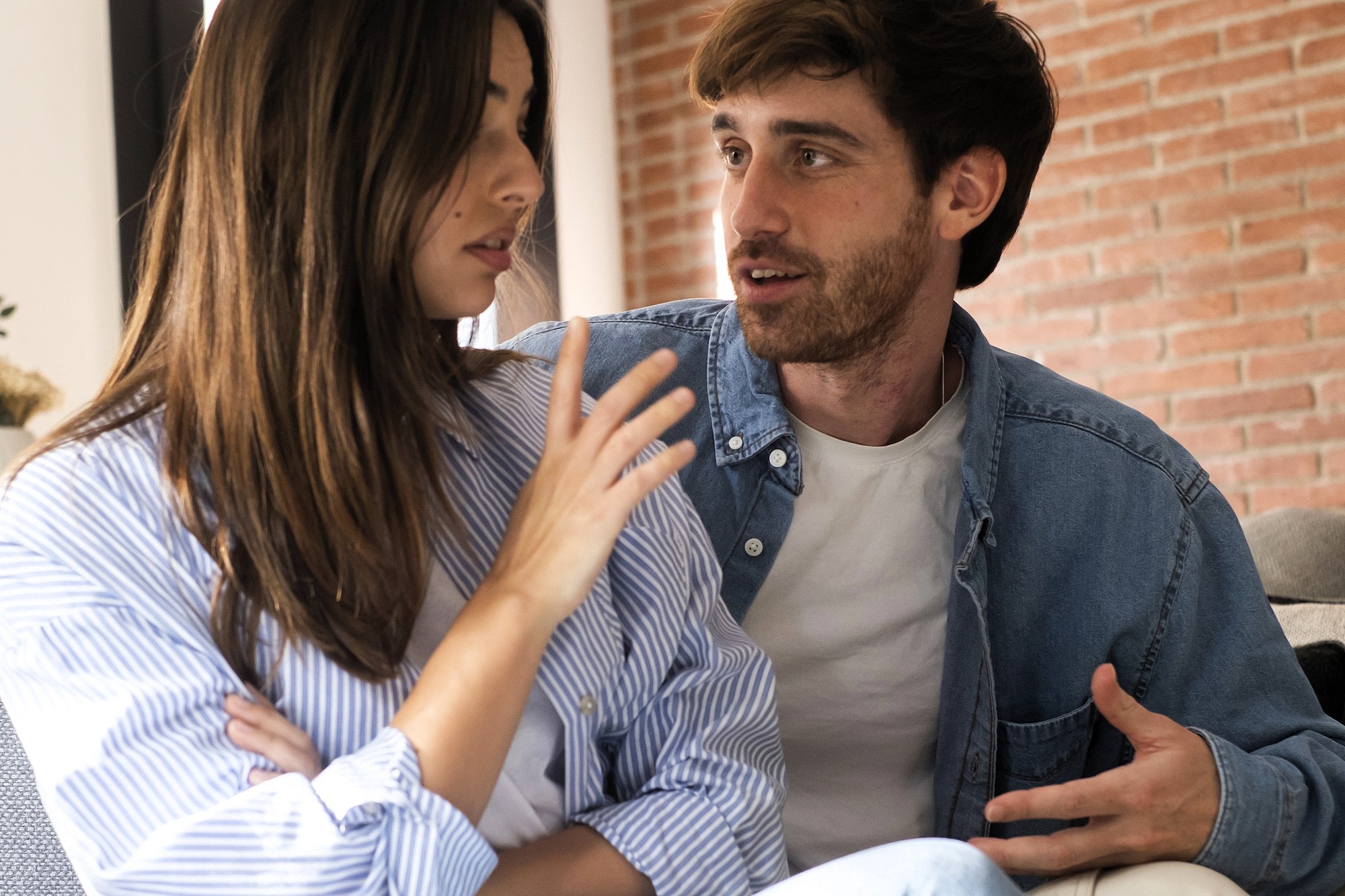 A woman and a man sit close together having a serious conversation. The woman looks away with her arms crossed while the man gestures with his hands, speaking earnestly. They are indoors with a brick wall behind them.