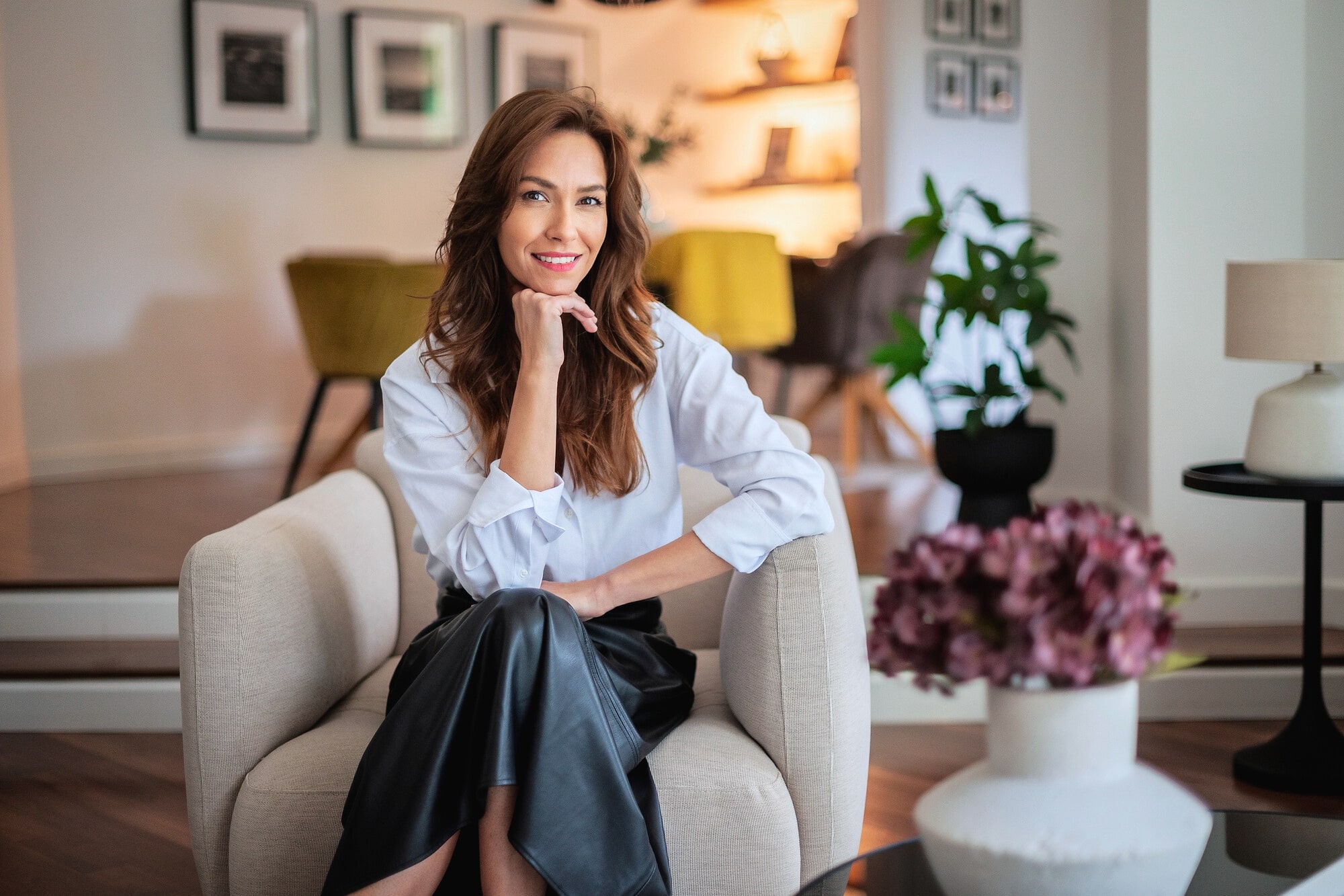 A woman with long brown hair, wearing a white blouse and black skirt, sits smiling in a modern, stylish living room with plants, flowers, and framed photos on the wall.